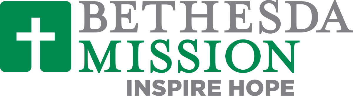 A middle-aged man in a blue sweater and beige pants sitting on a cushioned bench against a plain beige wall, with his hands resting on the bench and looking to the side.