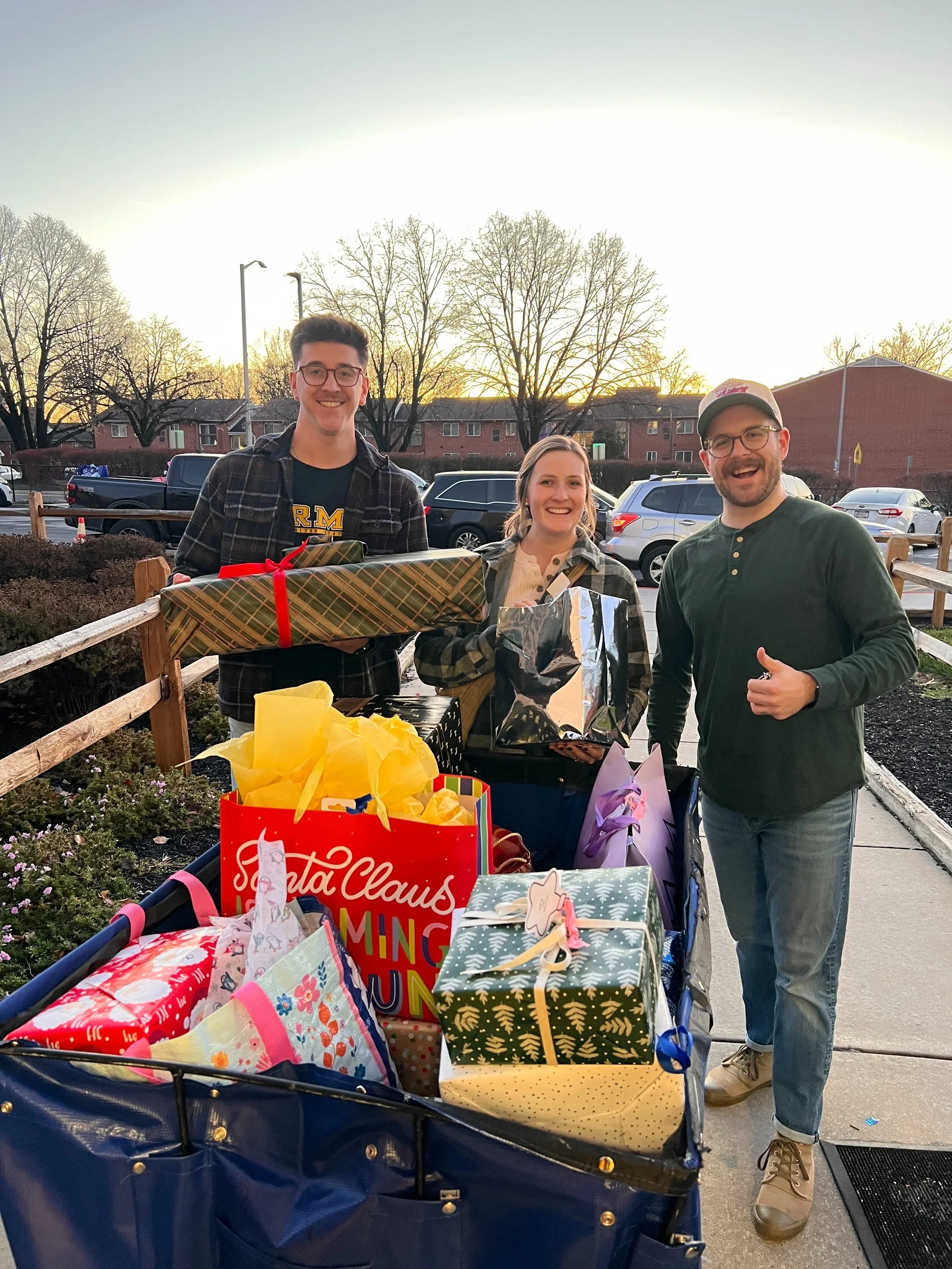 Three people standing outside with a wagon filled with Christmas gifts, smiling. It is sunset, and trees and parked cars are in the background.