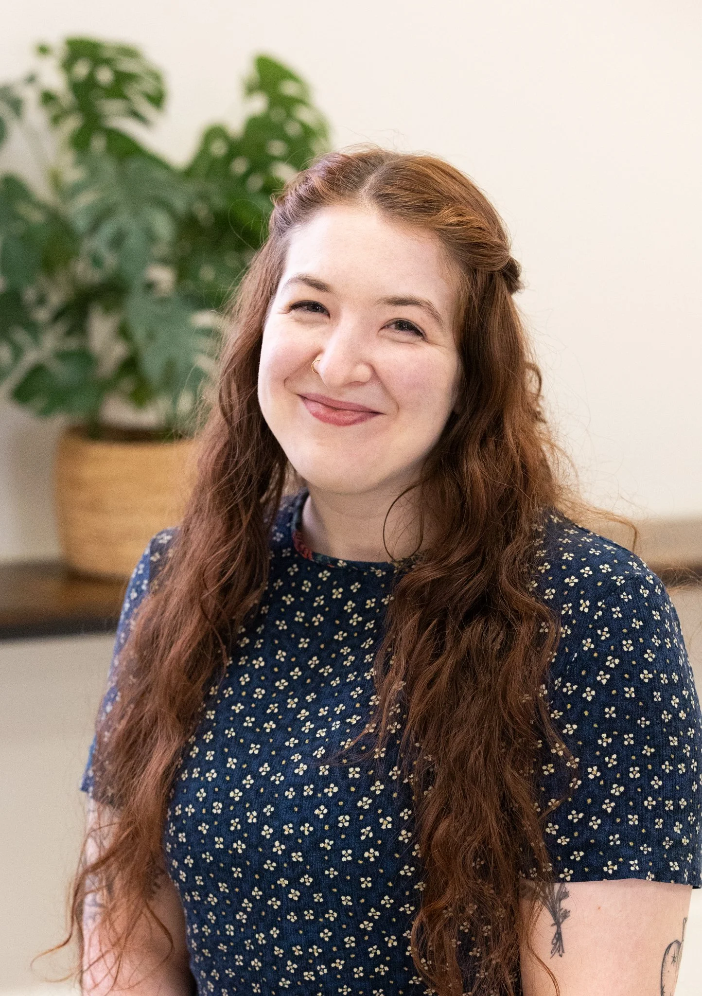 A woman with long, curly red hair smiling, wearing a dark blue dress with a small floral pattern, standing indoors with a green leafy plant in a basket behind her.