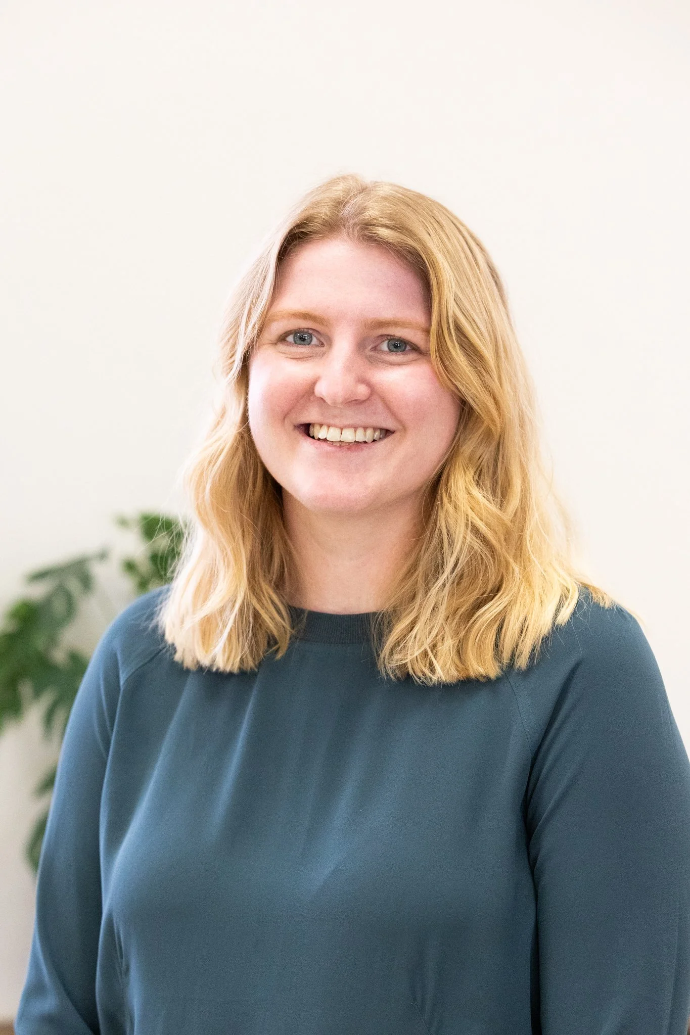 A woman with blonde, shoulder-length hair, smiling, wearing a dark teal top, standing in front of a pale background with a green plant in the background.