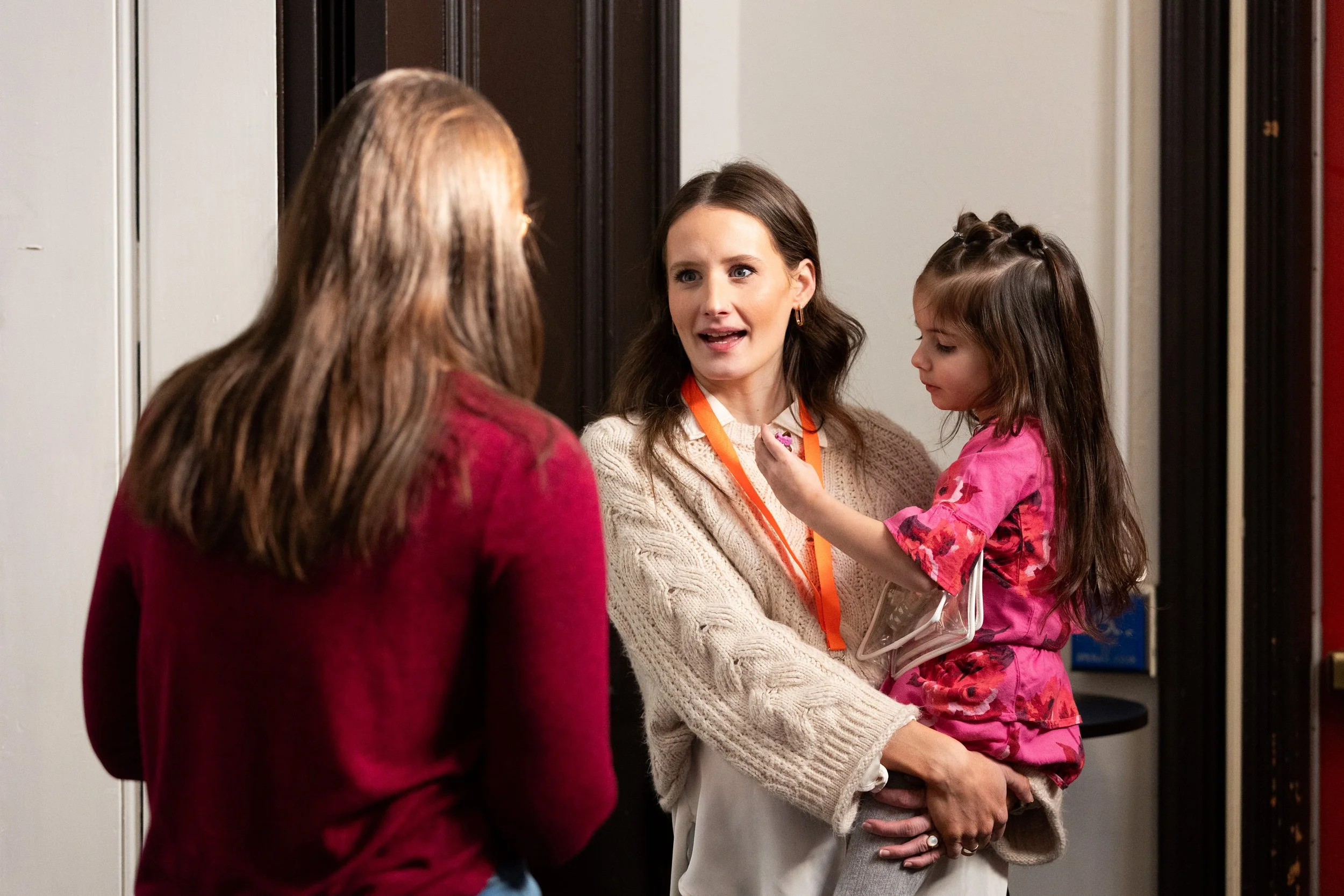 A woman holding a young girl in her arms, talking to another woman. The woman with a young girl is wearing a cream-colored sweater and a lanyard, while the girl is wearing a pink outfit. The woman with her back to the camera is wearing a red sweater. They are standing in front of a door.