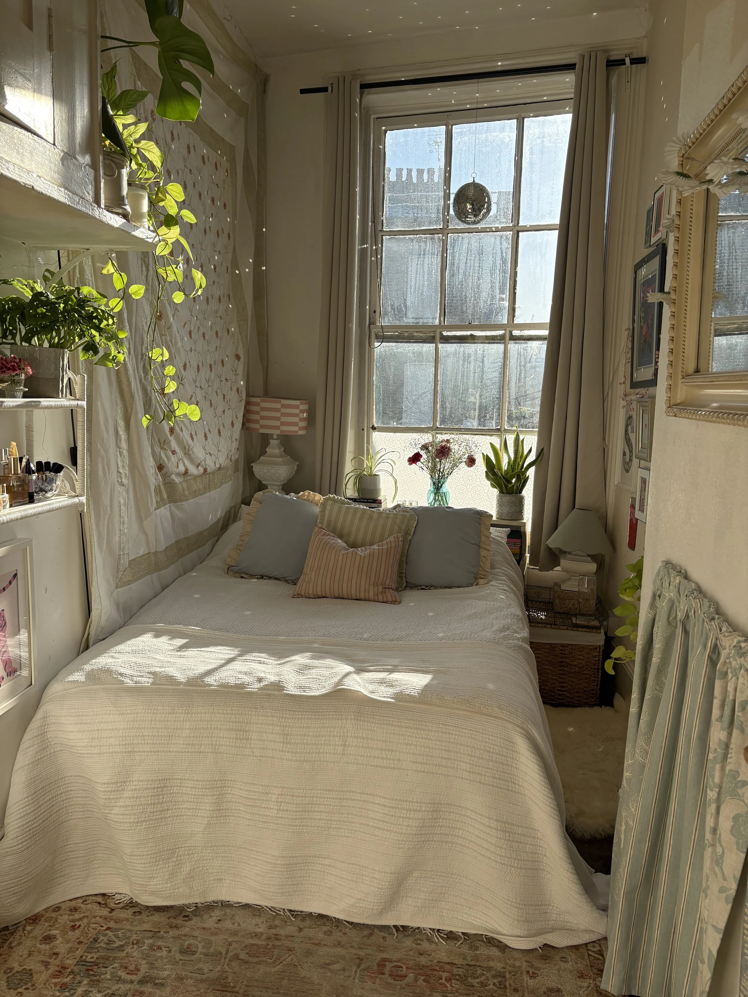 A cozy bedroom with a large window, sunlight streaming in, and various plants on the windowsill and shelves. The bed has a white cover with pillows, and there are decorative items and framed pictures on the walls.