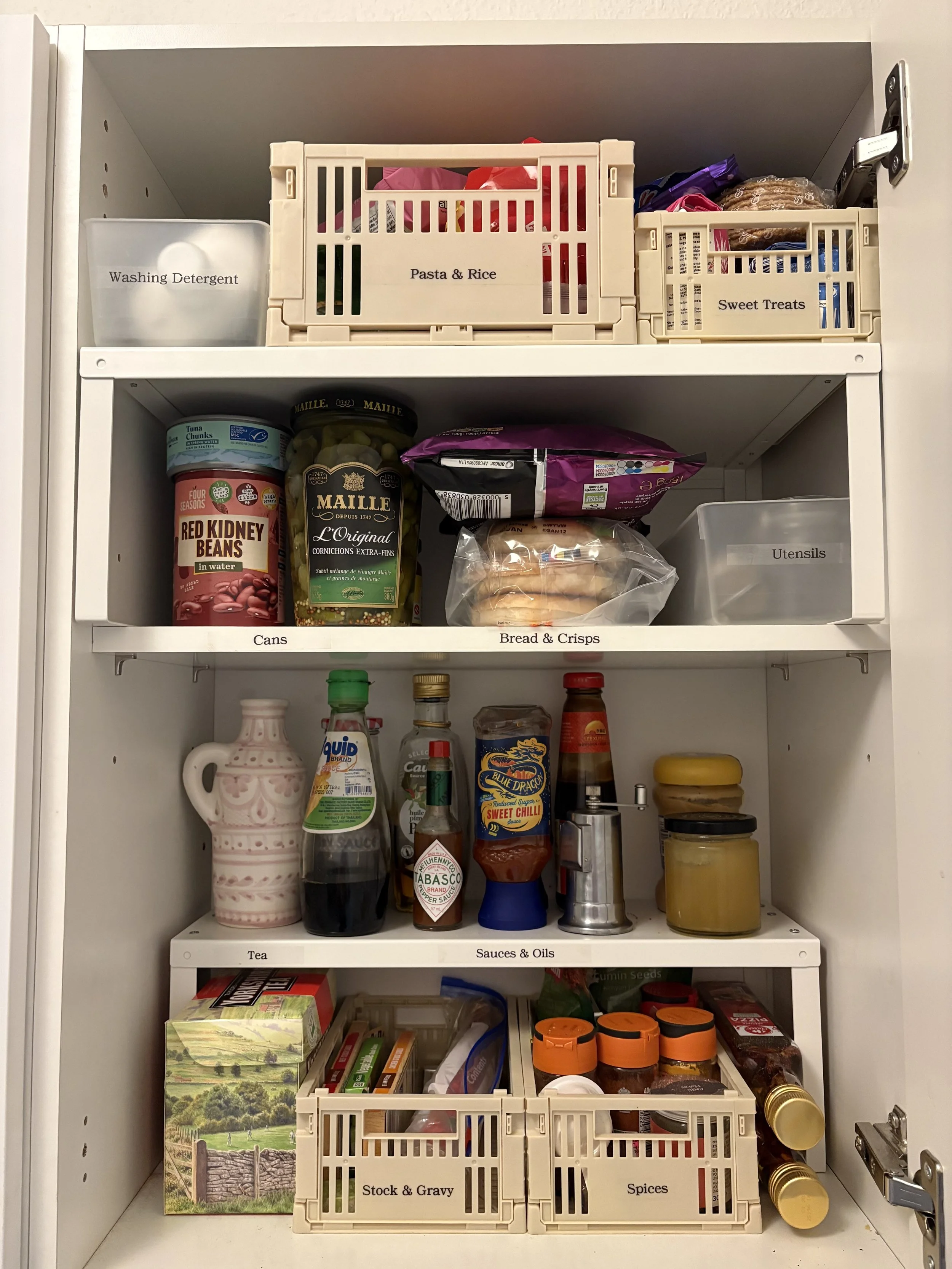 Open kitchen cabinet with labeled compartments containing pantry items such as canned goods, sauces, spices, bread, treats, utensils, and cleaning supplies.