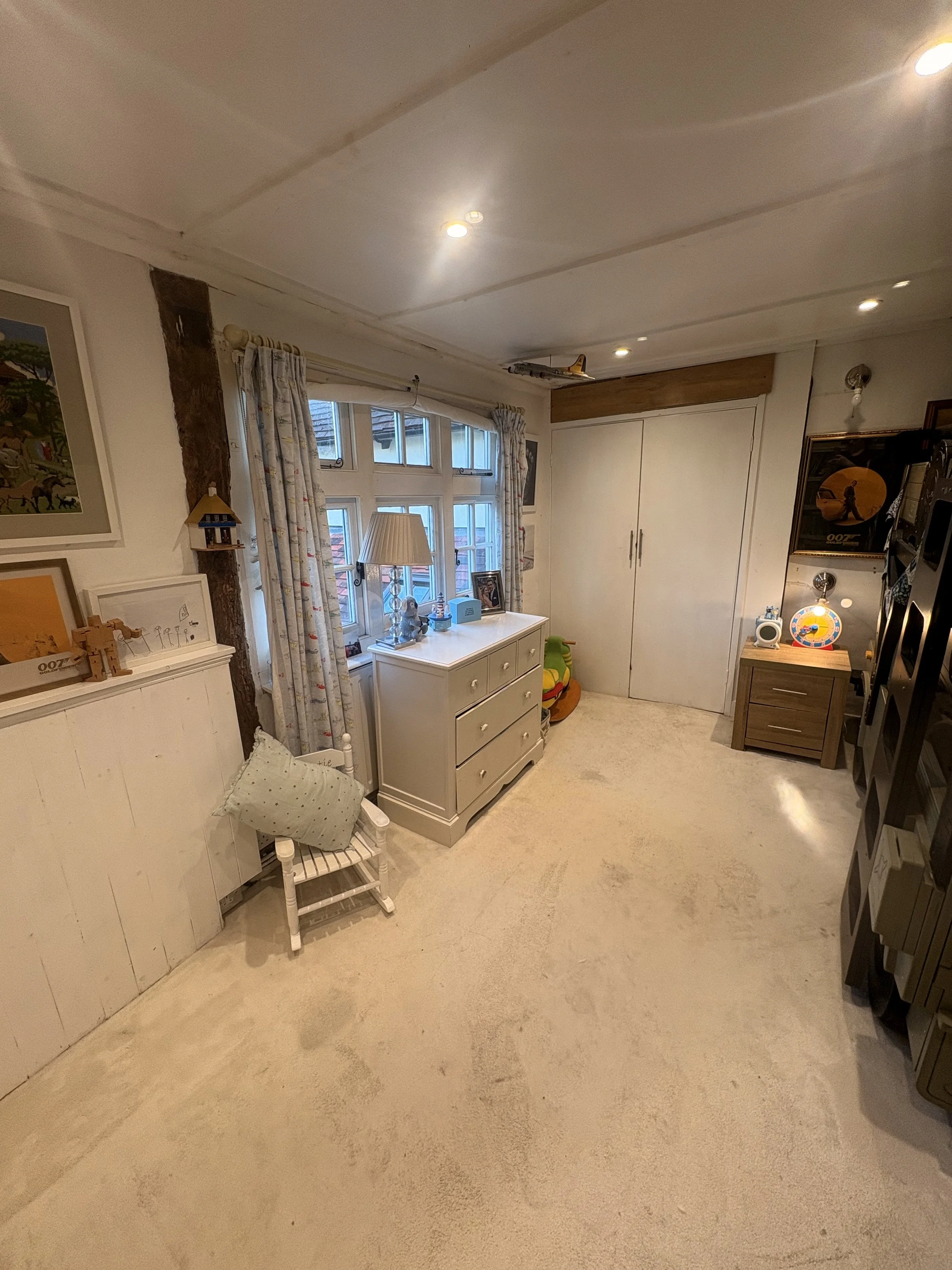 A cozy bedroom with white walls and beige carpet. A window with floral curtains, a small white chair with a pillow, a white dresser with a lamp and decorative items, and some framed pictures on the wall.
