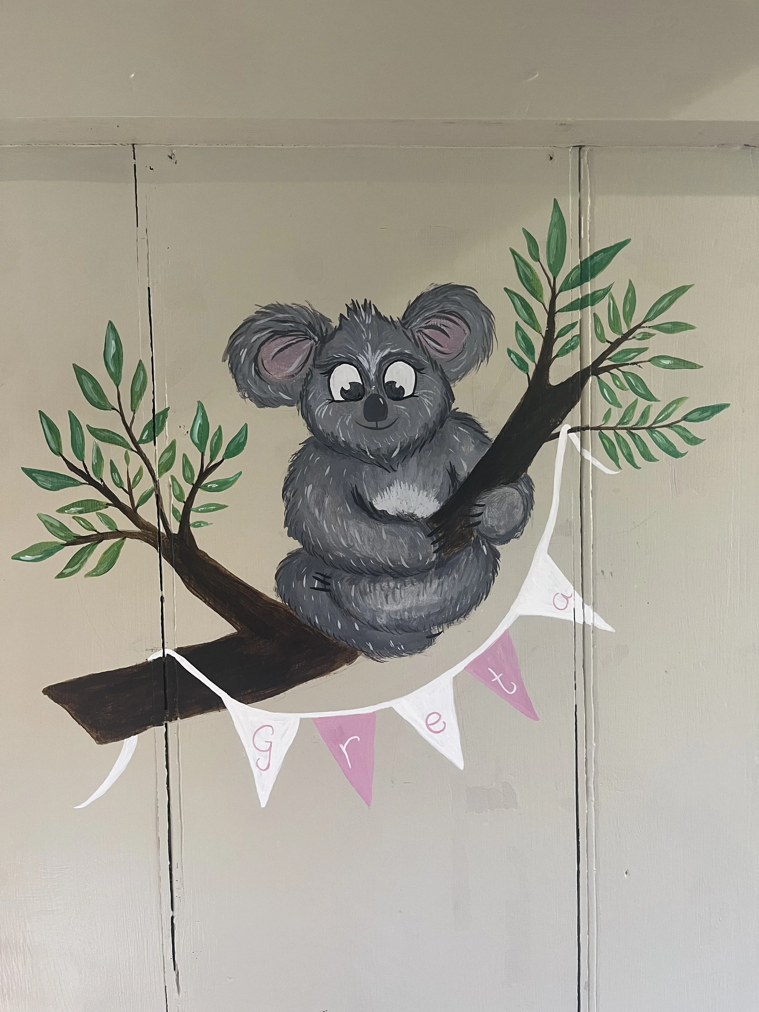A painting of a cute gray koala bear with big ears sitting on a painted tree branch, holding onto the branch with its paws, and a banner hanging below with the name 'Grett' in pink and white flags.