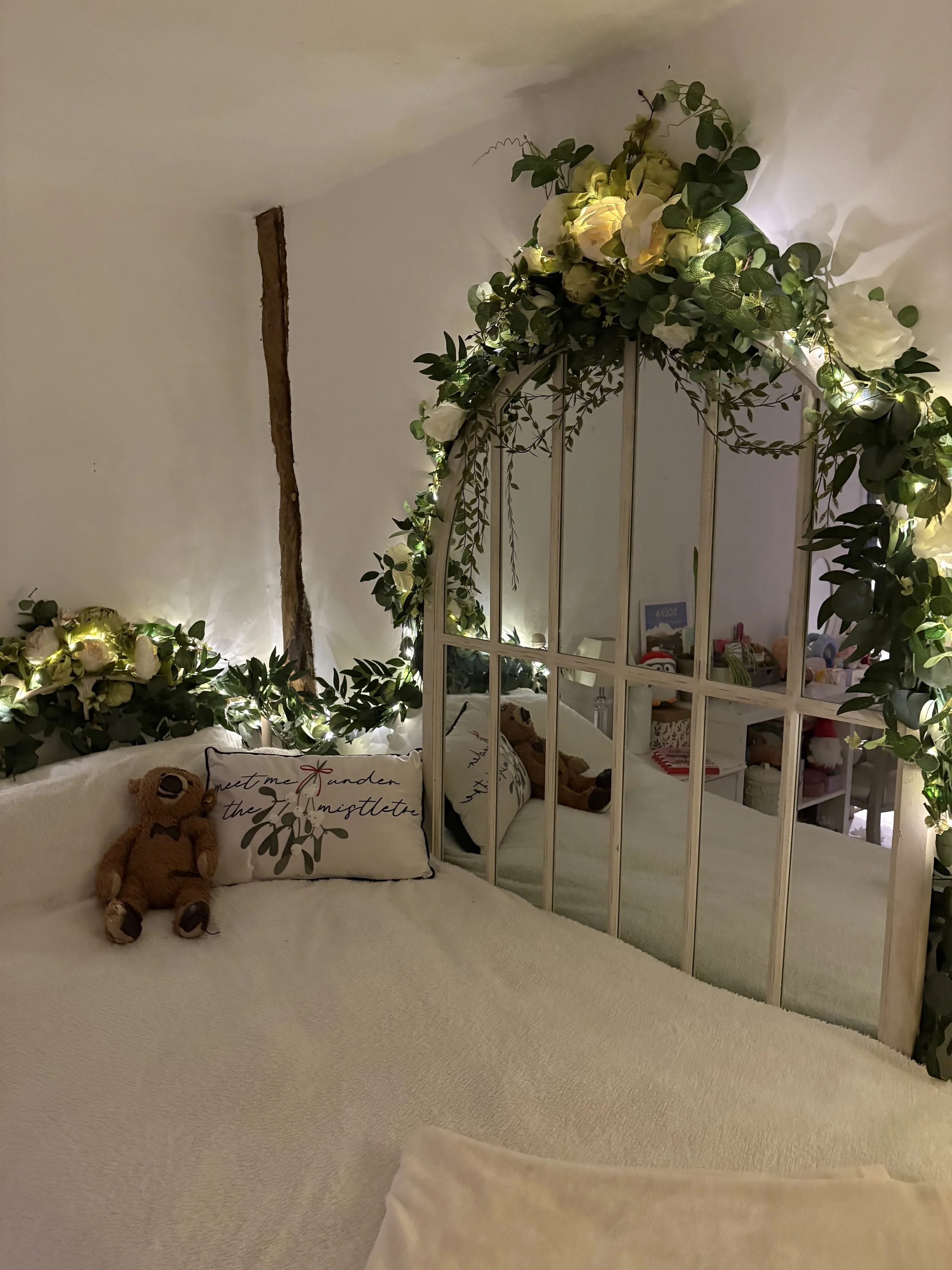 A bedroom decorated with string lights, green foliage, and white flowers forming an arch above a mirrored headboard. There are pillows with writing and stuffed bears on the bed.