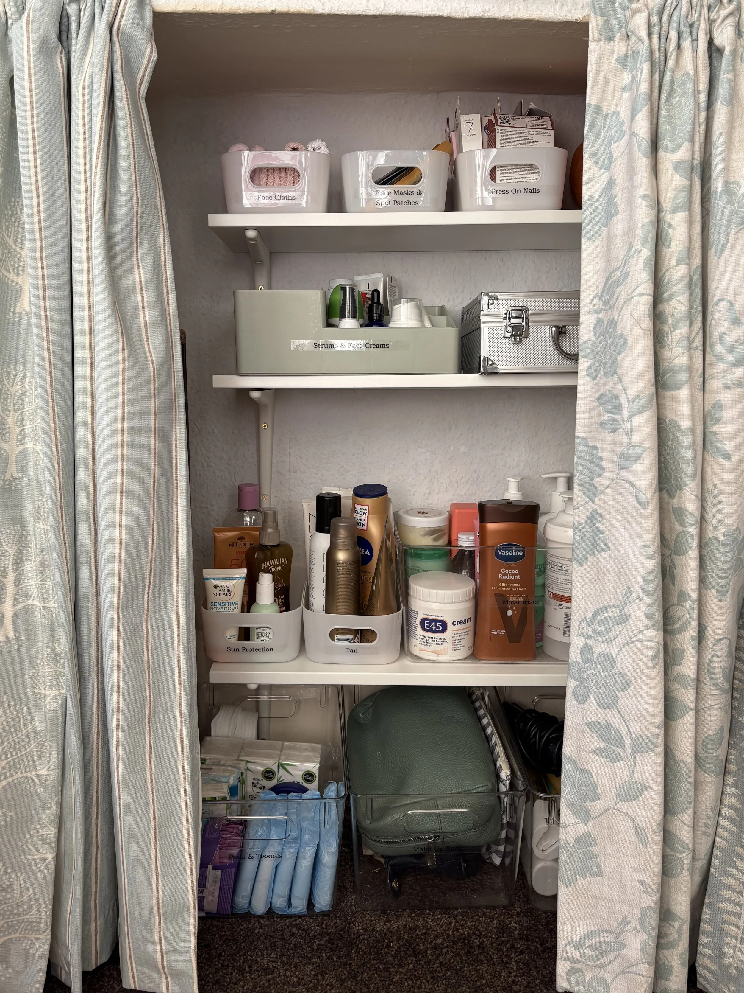 A closet with three white shelves stocked with personal care products, creams, and toiletries. The bottom shelf has organized beauty and skincare items, with tissues and makeup bags in clear containers. The closet is partially covered with patterned curtains on either side.