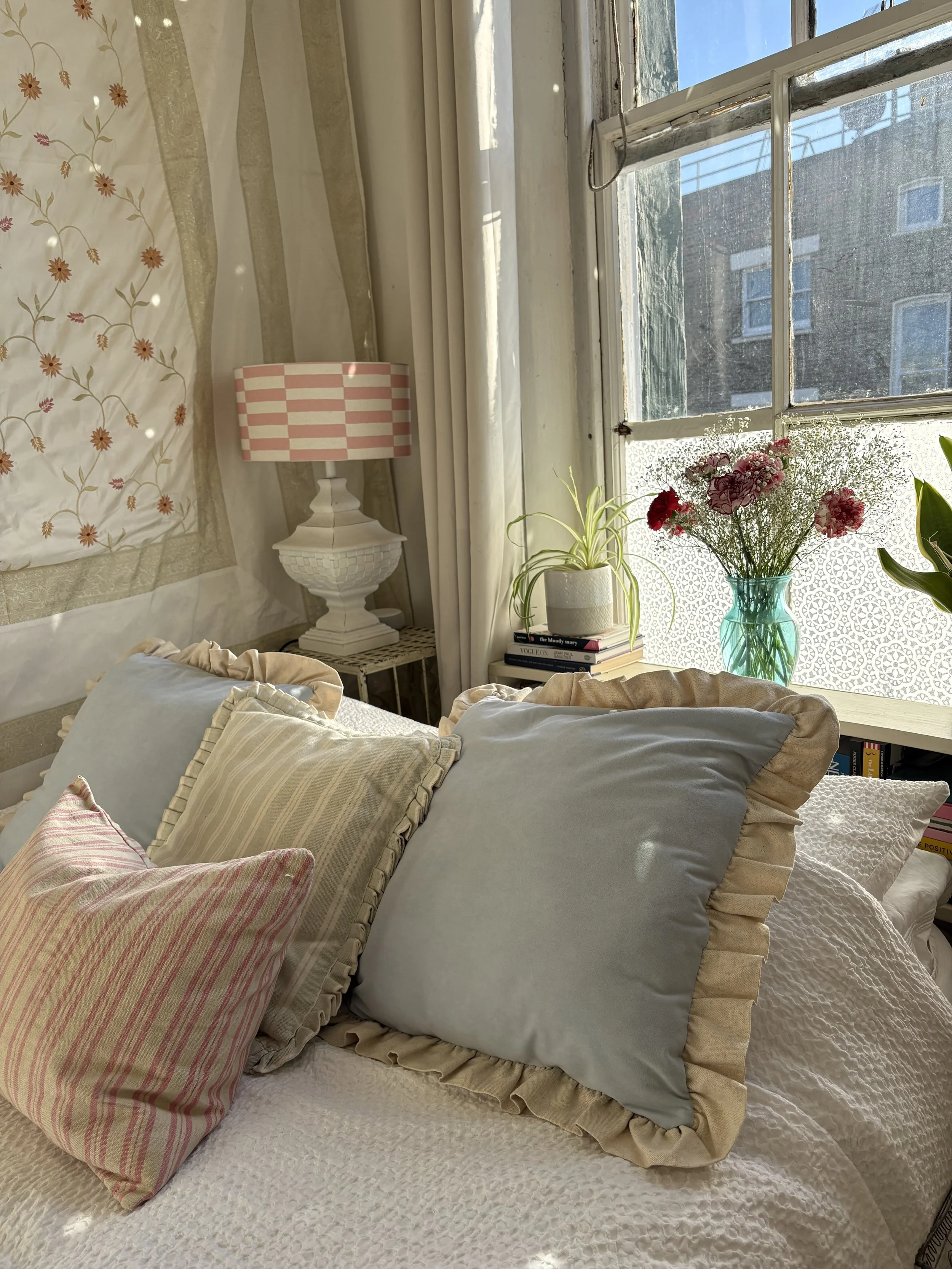 A cozy bedroom with a bed covered in white textured bedding, decorated with three pillows in pastel shades with ruffled edges. A side table holds a pink and white checkered lamp and a vase of pink and white flowers, with a window and plants in the background.