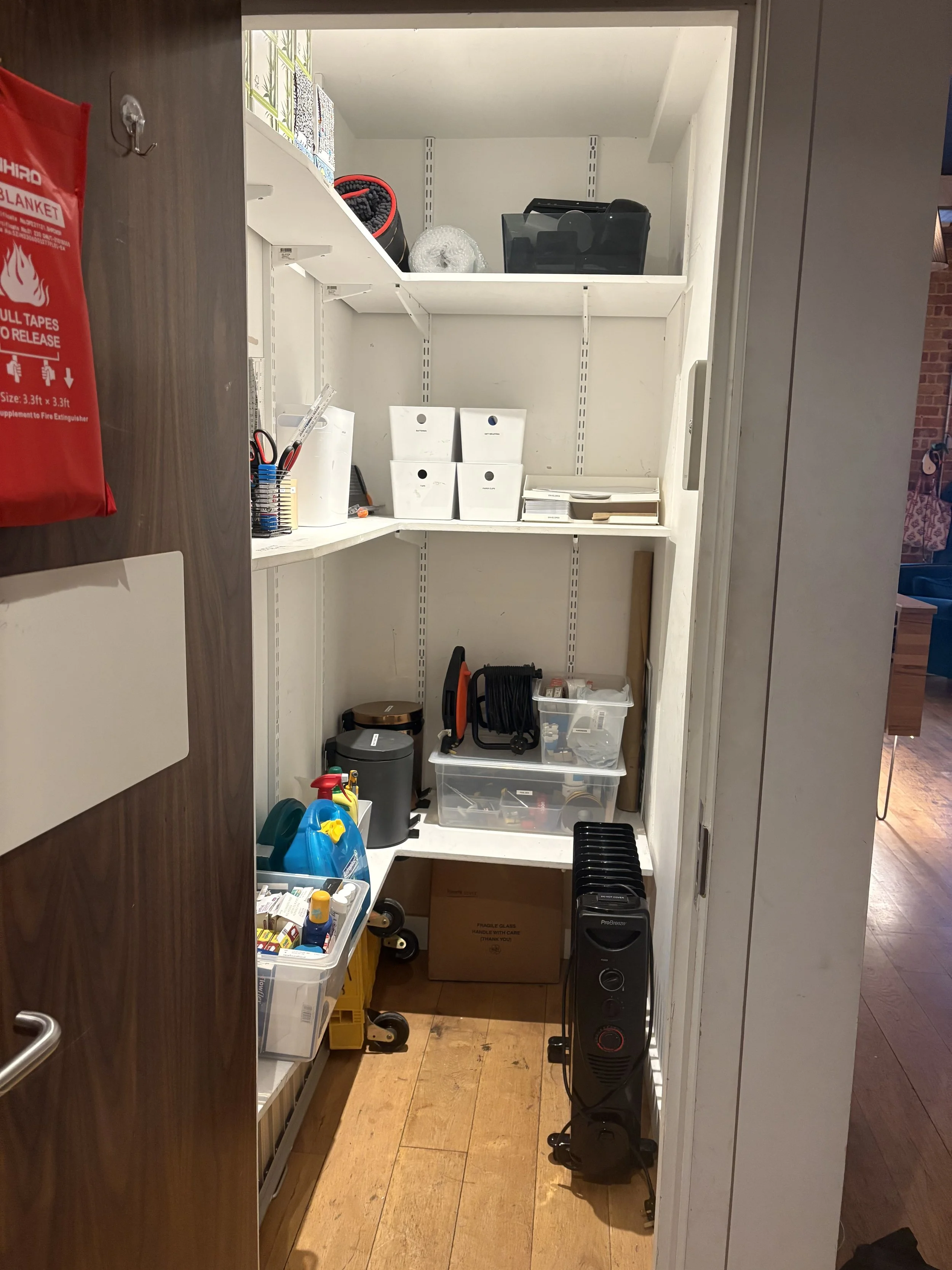 A small storage closet with shelves containing various cleaning supplies and tools, including spray bottles, a vacuum cleaner, and plastic containers.