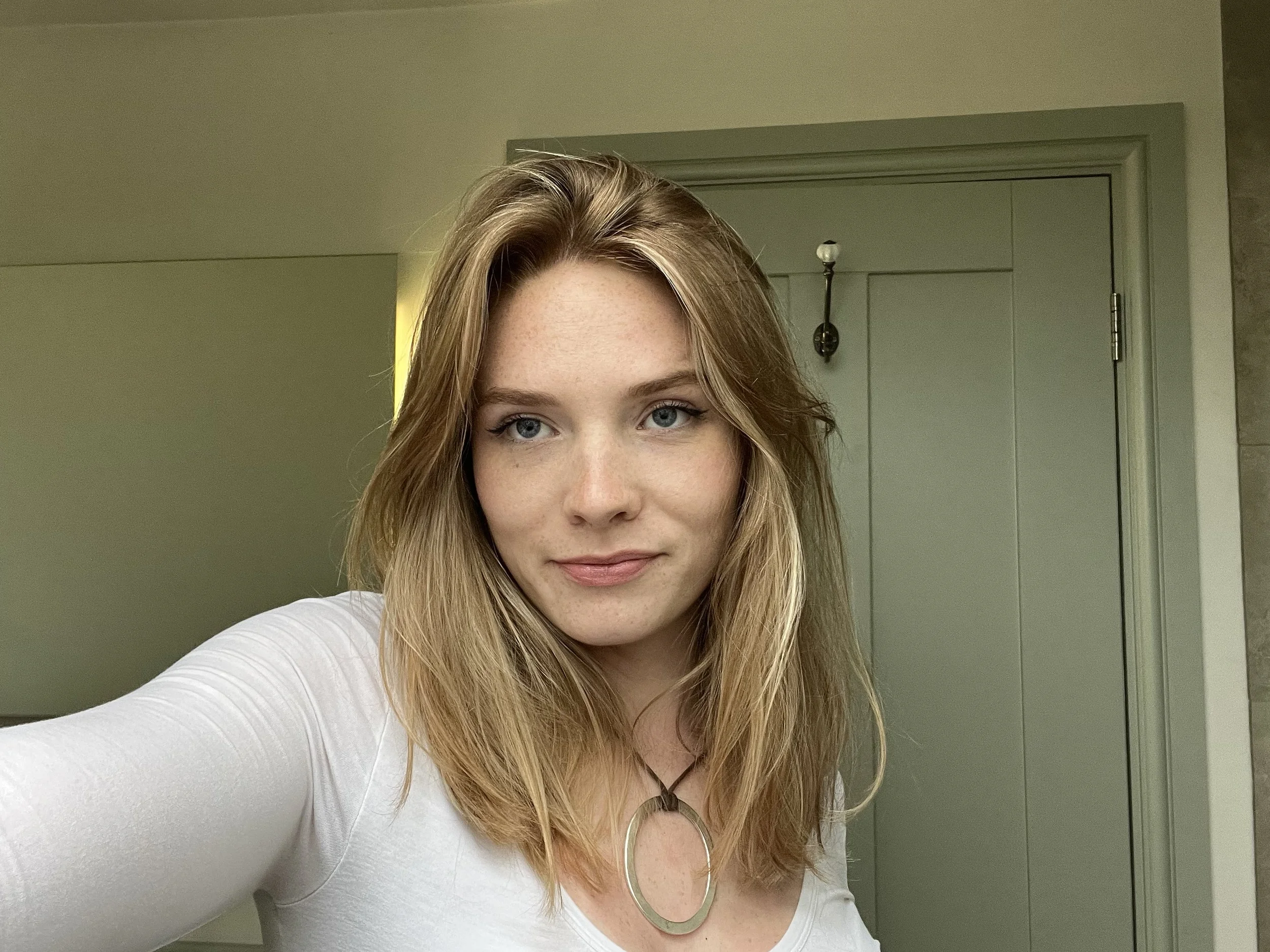 A young woman with blonde hair and blue eyes taking a selfie indoors. She is wearing a white top and a circular pendant necklace, with a calm expression on her face.