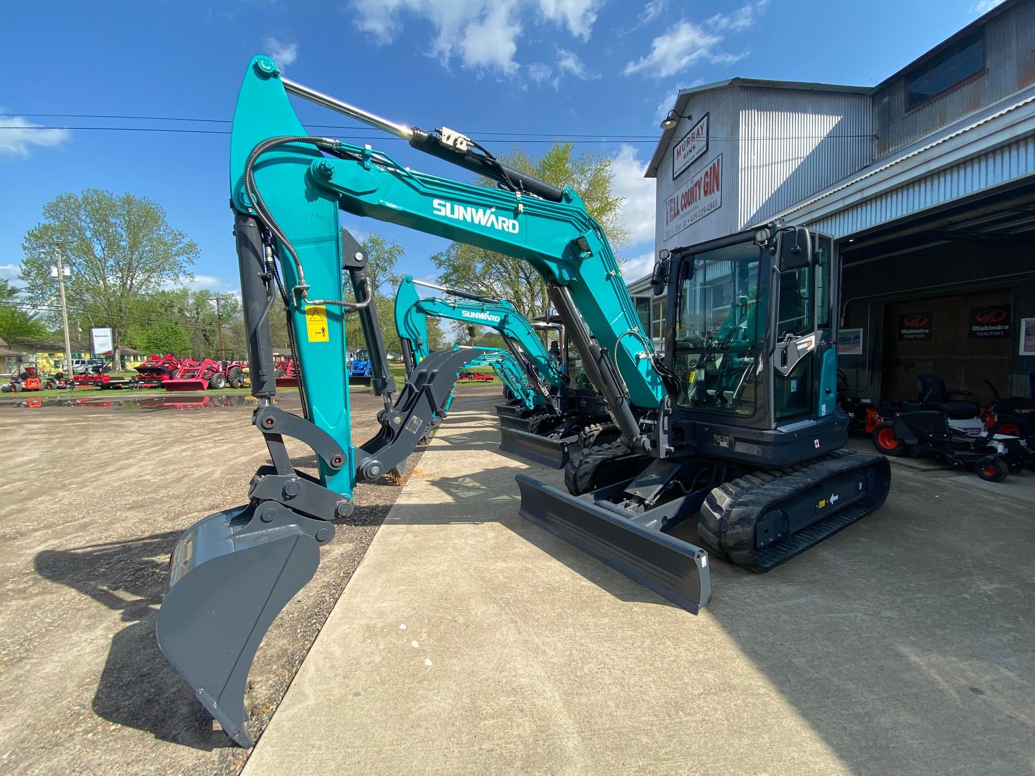Blue Sunward compact excavator with a bucket attachment parked outside Yell County Gin Co.  with other machinery and equipment visible in the background.