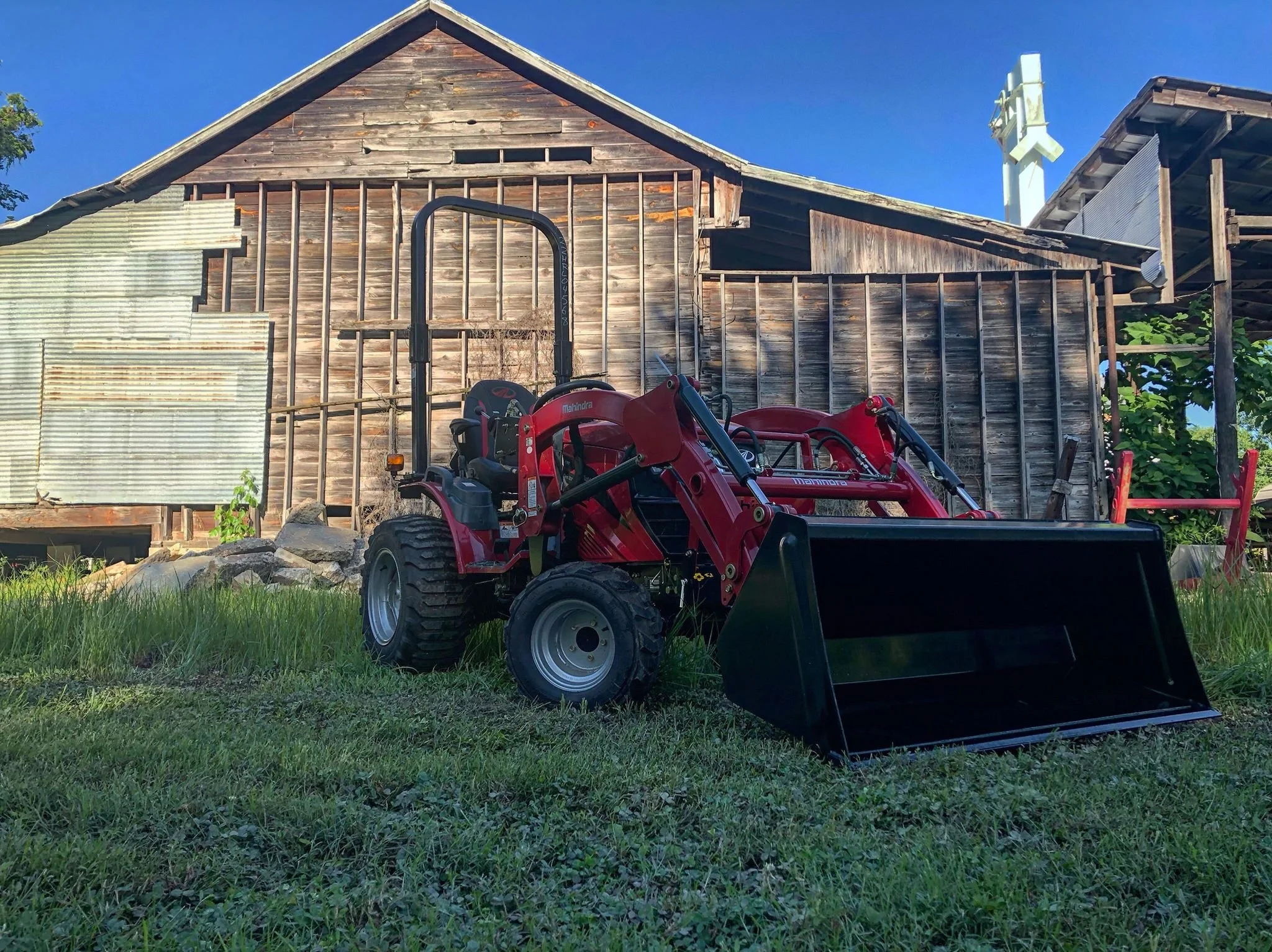 Red mahindra compact utility tractor with front loader attachment parked on grass in front of a weathered wooden barn. located at Yell County Gin Co. in Dardanelle, Arkansas