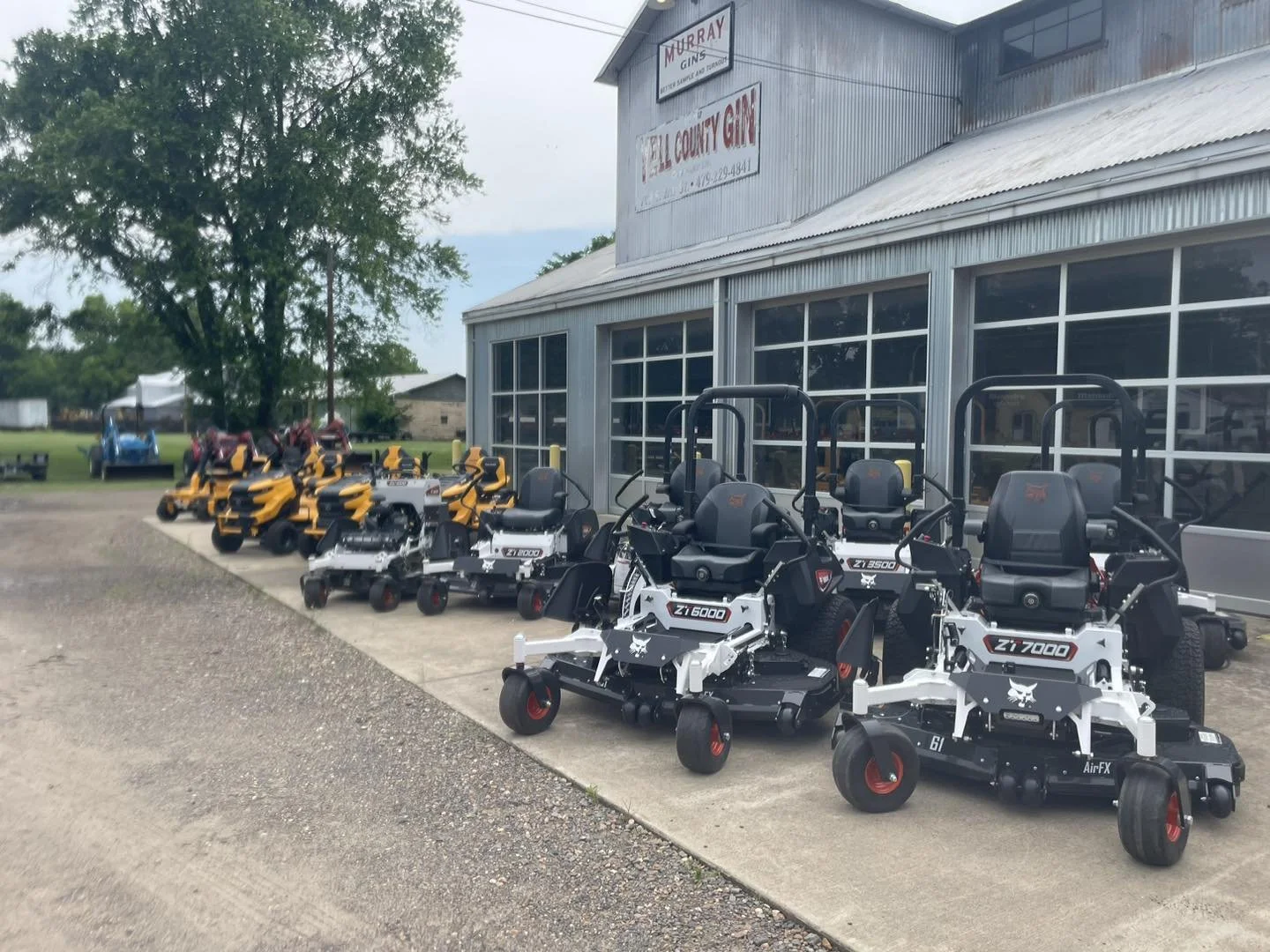 Multiple bobcat and hustler lawn mowers of various sizes and colors, including black, white, yellow, and blue, lined up outside of Yell county gin co.,  a metal building with large windows and signage for Murray Gin and Yell County Gin.