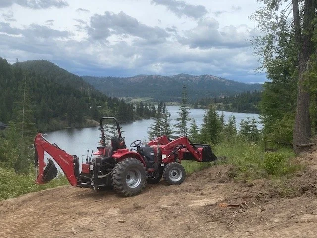 A red mahindra 1626 tractor with a front loader attachment parked on a dirt area near a lake, surrounded by trees and mountains under a cloudy sky. located in rural Montana