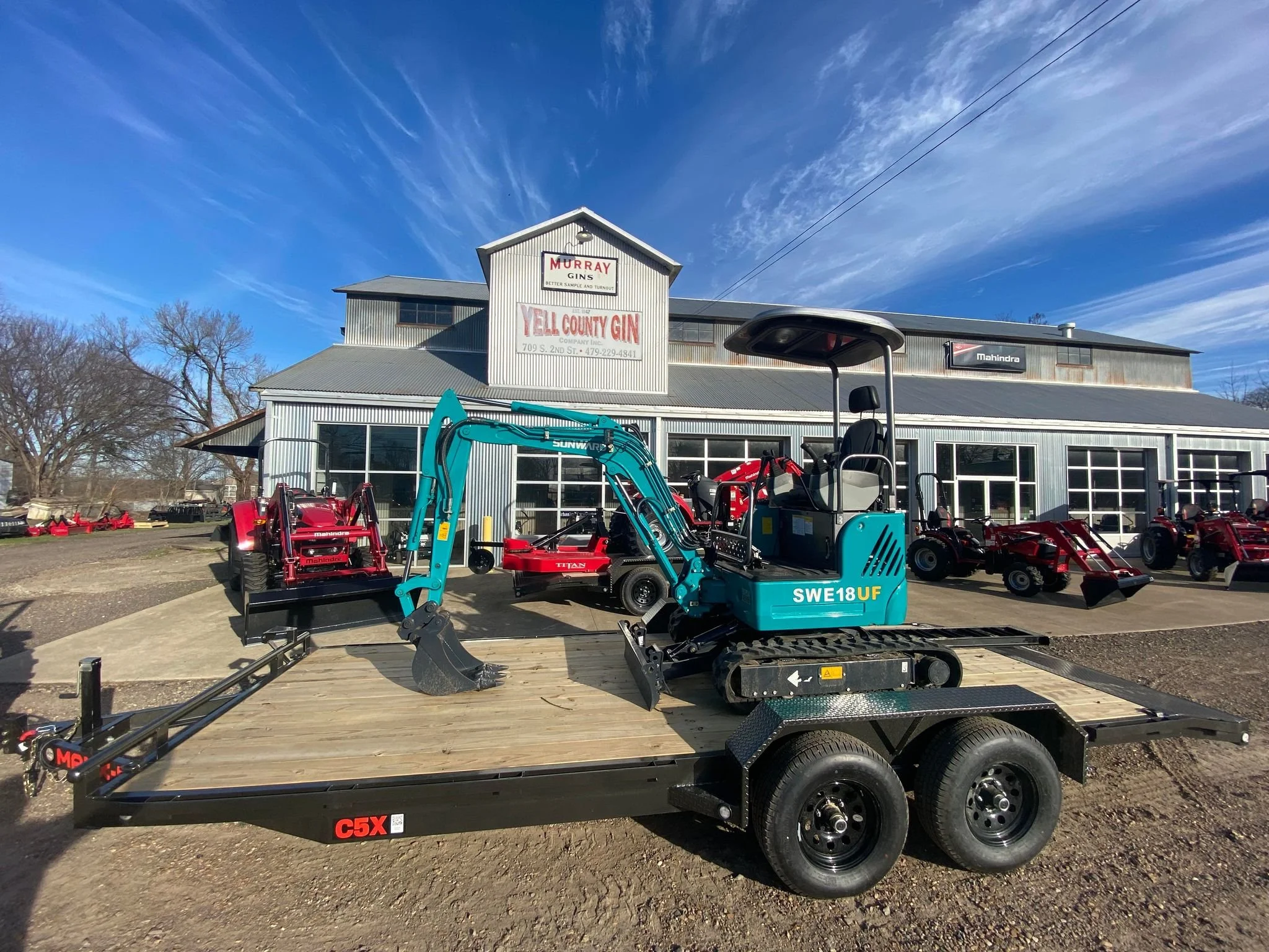 A Sunward mini excavator on a black trailer in front of a gray industrial building with signs for a gun shop and gin store, and several red tractors or lawn equipment located nearby. Located at Yell County Gin Co. 
