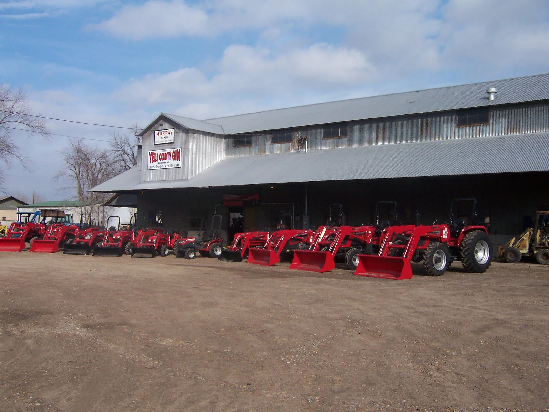 Red tractors parked in front of Yell County Gin, a retired cotton gin. The ground is dirt, and the sky is partly cloudy.