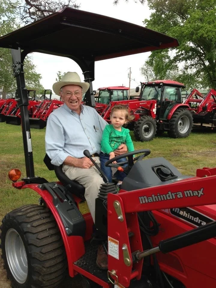 An elderly man in a white cowboy hat and light-colored shirt sitting on a red Mahindra Max tractor with a young in a green sweater sitting in his lap, both smiling. The background shows multiple red tractors parked on grassy ground under a cloudy sky, with trees and utility poles nearby.