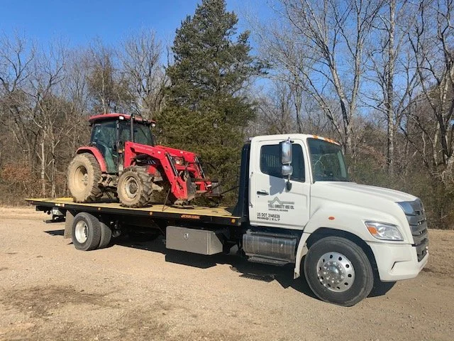 White flatbed truck from Yell County Gin Co. carrying a red Mahindra tractor on a dirt road with leafless trees and a blue sky in the background.