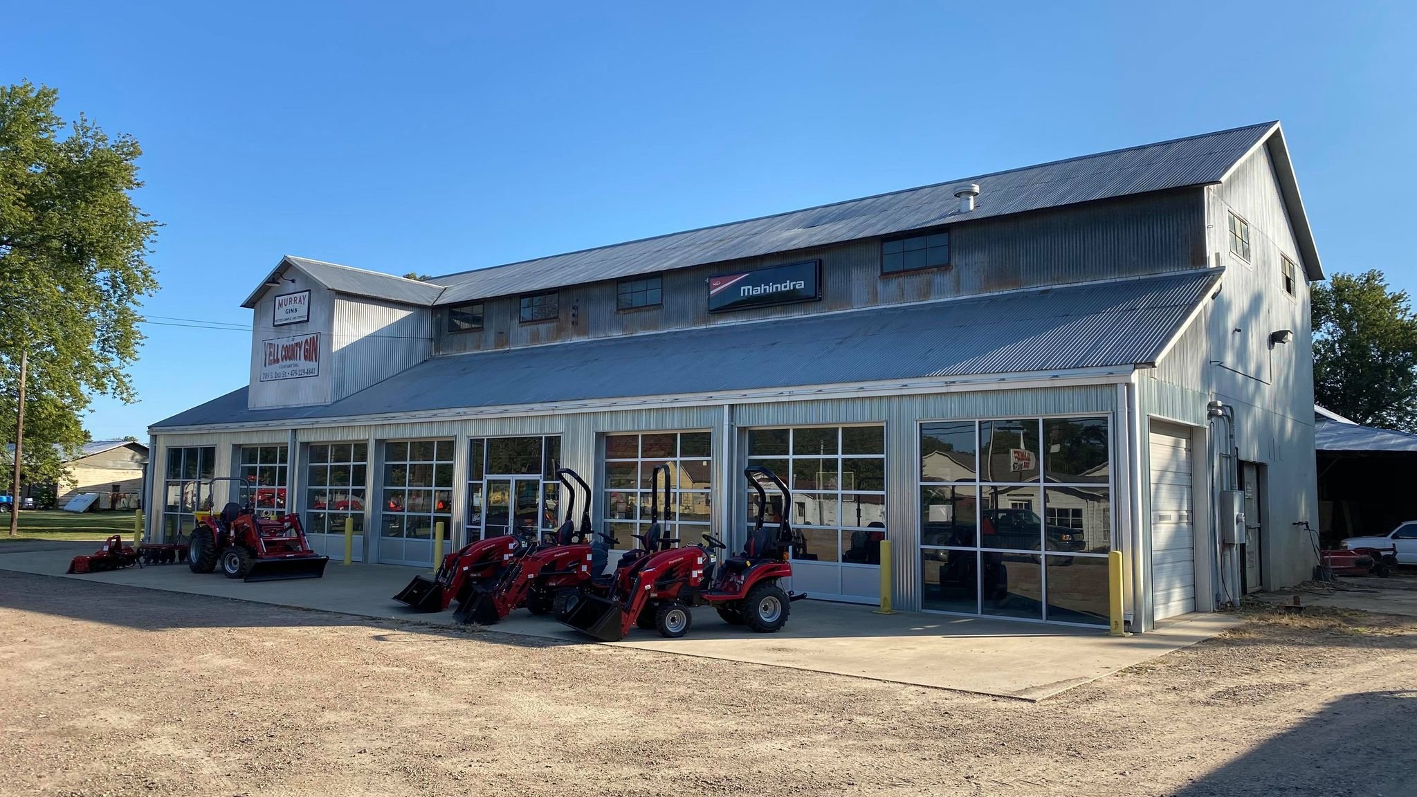 A recent picture of Yell County Gin Co., a large retired cotton gin with large glass windows, and signs including "Murray Gins" and "Mahindra," with red riding lawnmowers and equipment outside on a concrete slab, against a bright blue sky.