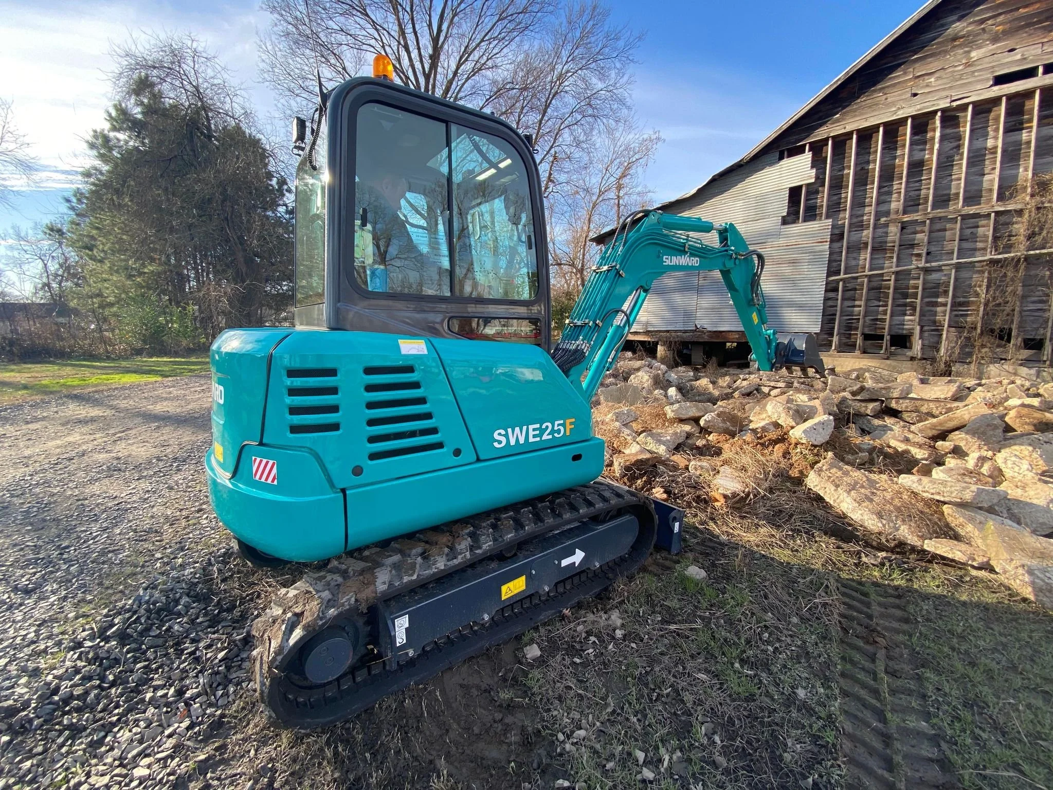 Small teal excavator working on a foundation with rocks near an old wooden building. Located at Yell County Gin Co. 