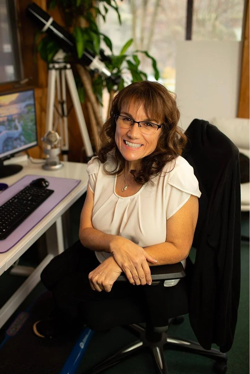 A woman with brown hair and glasses smiling at her desk, with a computer, a microphone, a telescope, and a large green plant in the background.