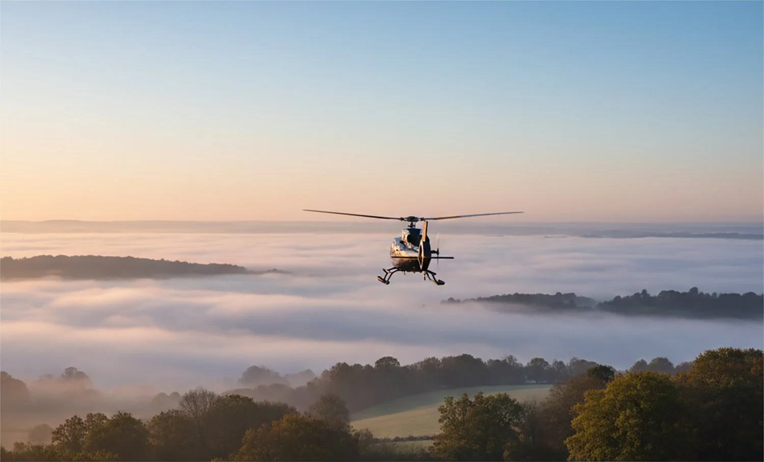 A helicopter flying above a landscape with trees, fog, and hills during sunrise or sunset.