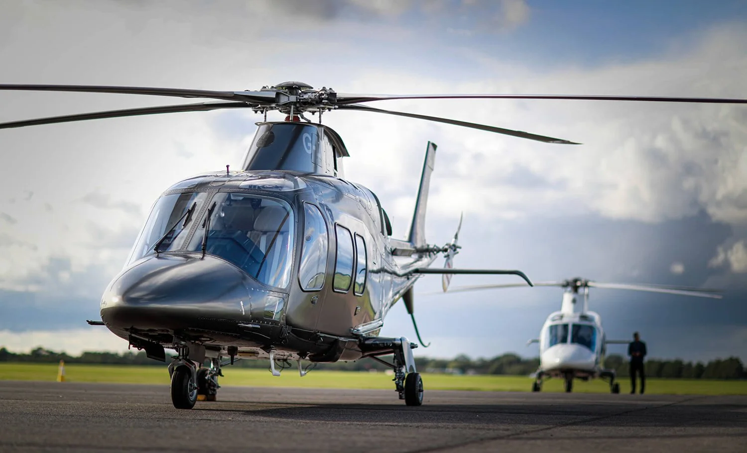 Two private charter helicopters on a runway with a person standing near the rear helicopter, under cloudy skies.
