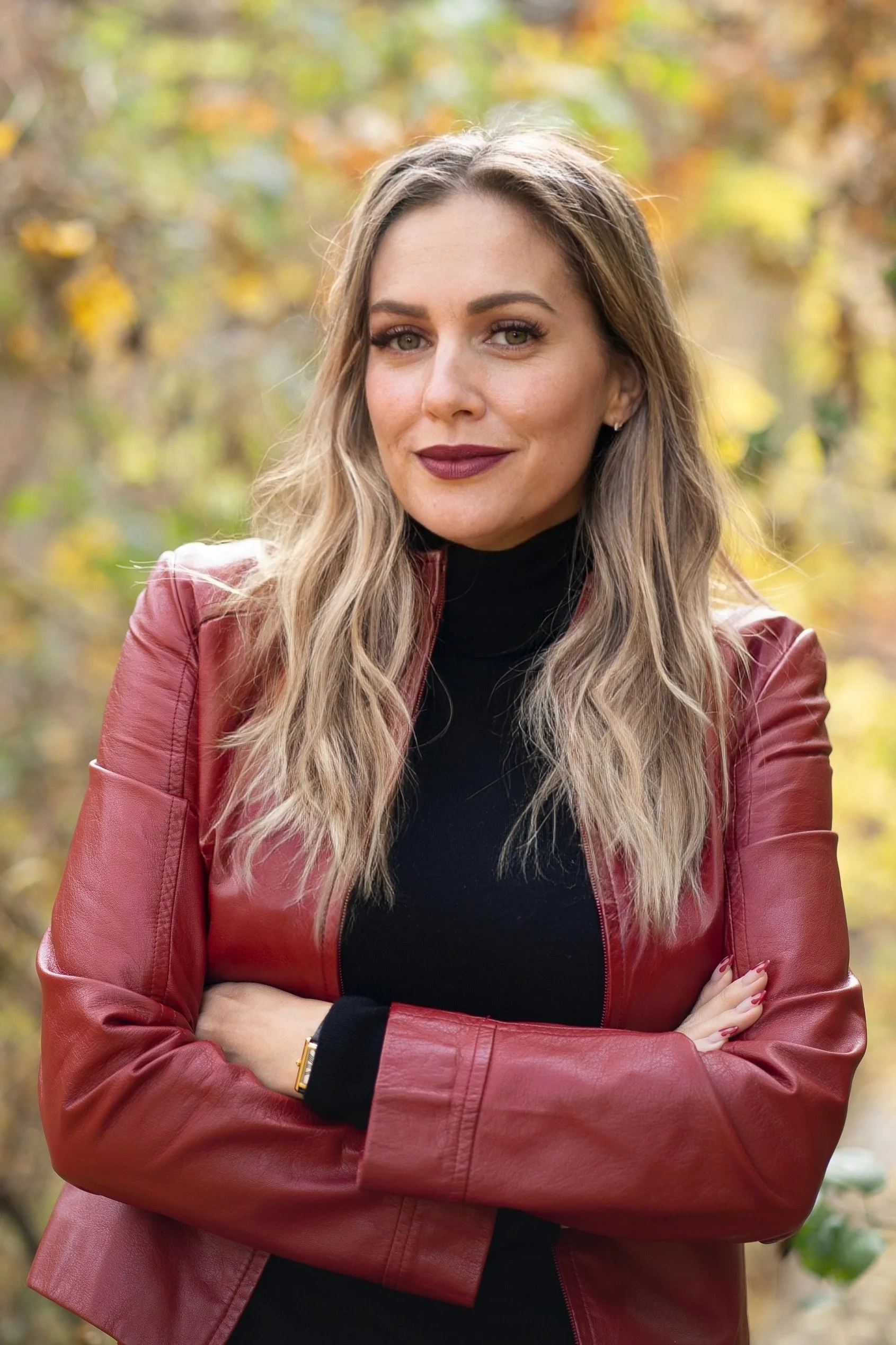 A woman with long, wavy blonde hair, wearing a red leather jacket over a black turtleneck, stands outdoors with arms crossed, smiling slightly, with autumn leaves in the background.