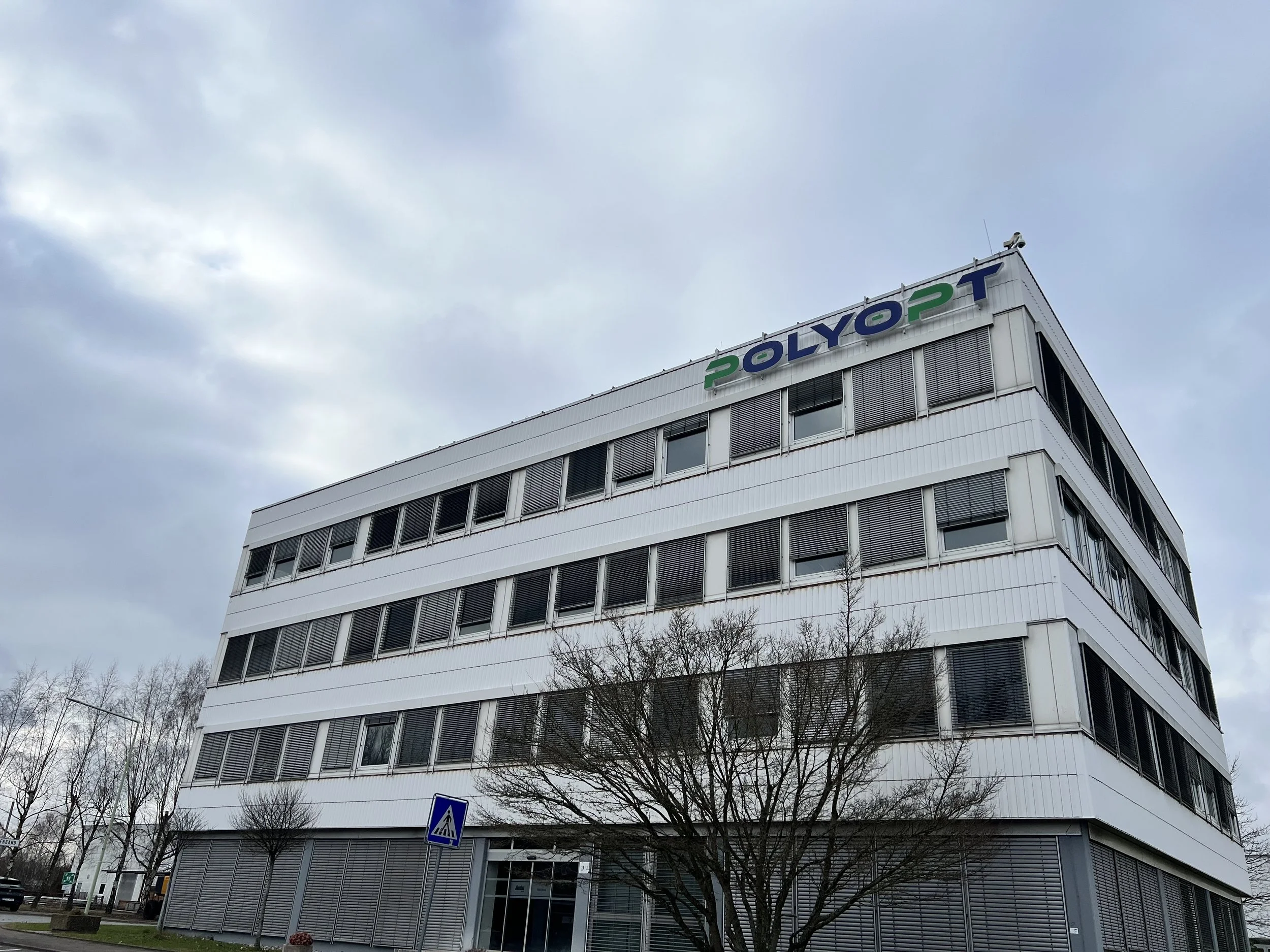 A white multi-story office building with a sign on the top that reads 'POLYOPT' in colorful letters, and a parking lot with a few trees and a pedestrian crossing sign in front.