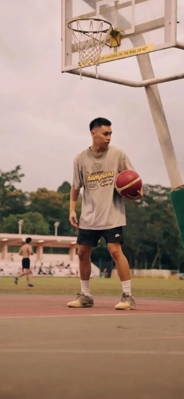 A young man holding a basketball standing on an outdoor basketball court near the hoop with a field and trees in the background.