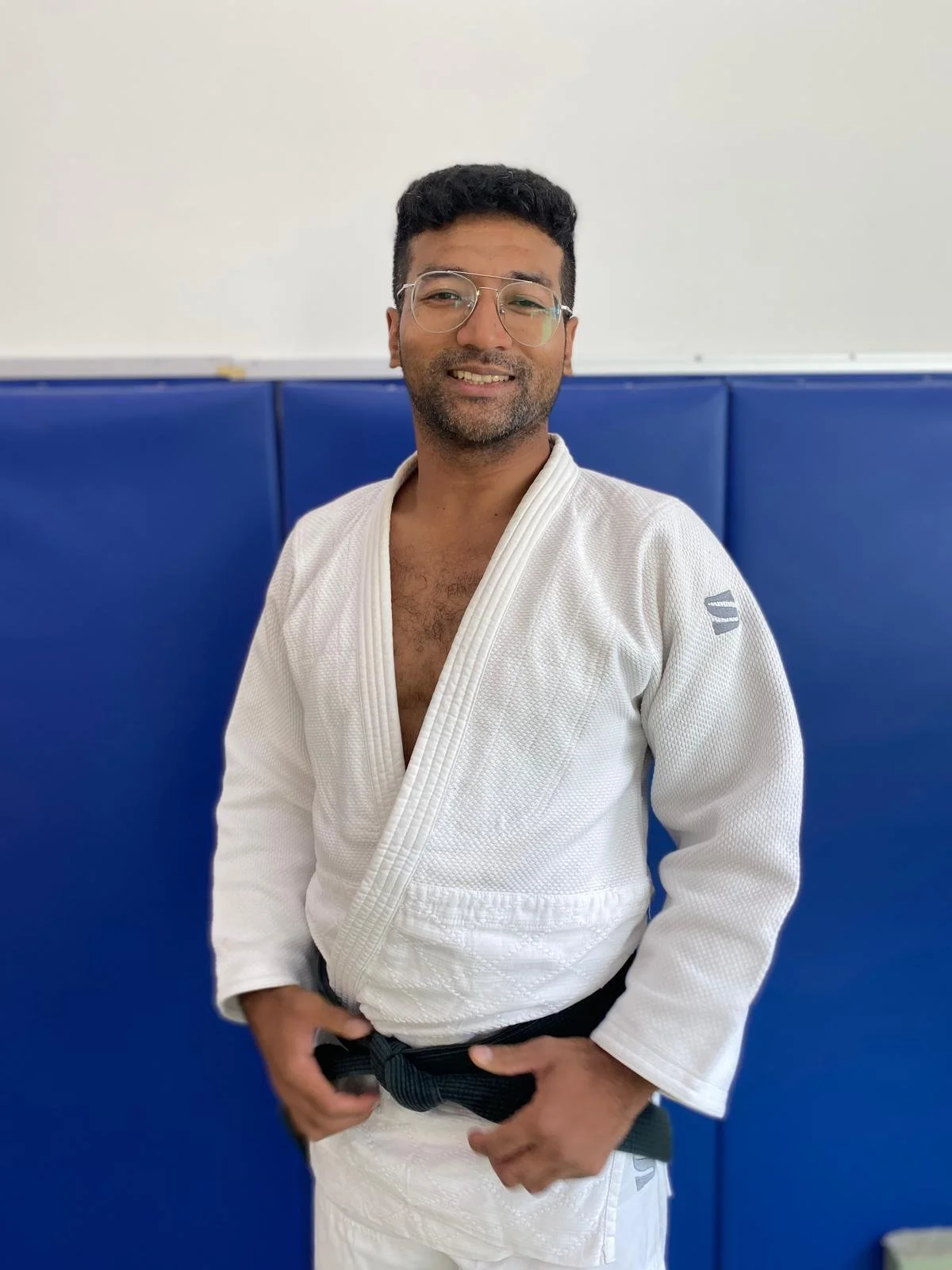A man in a white judo gi with a black belt, standing in front of blue padded walls, smiling at the camera.