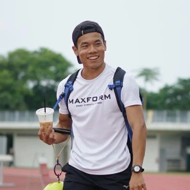 Young man smiling, wearing a black cap backwards, white t-shirt with 'MAXFORM' logo, carrying a blue backpack, holding a drink, standing outdoors on a track field with green trees in the background.