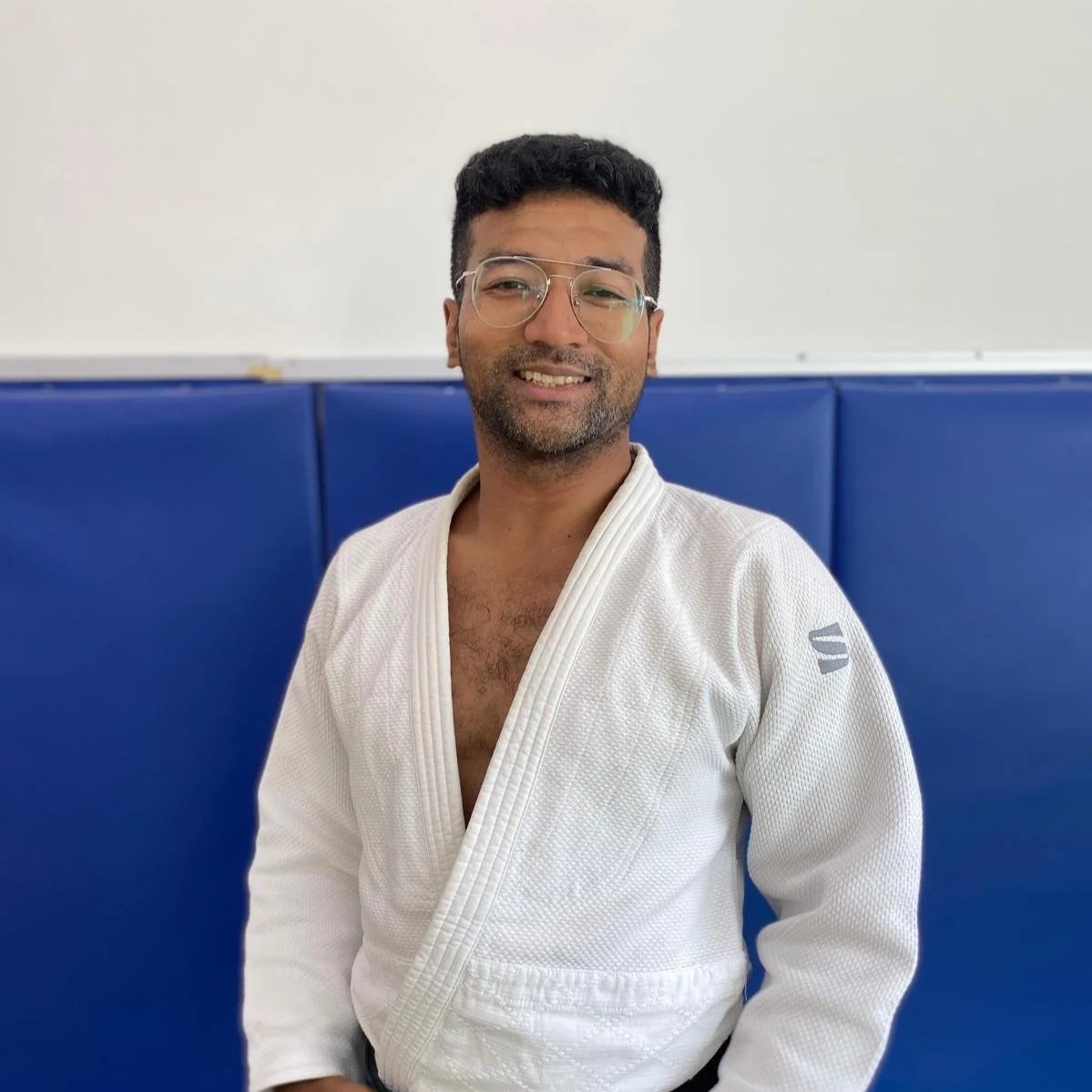 A man wearing glasses and a white judo gi, standing in a martial arts dojo with blue padded walls, smiling at the camera.