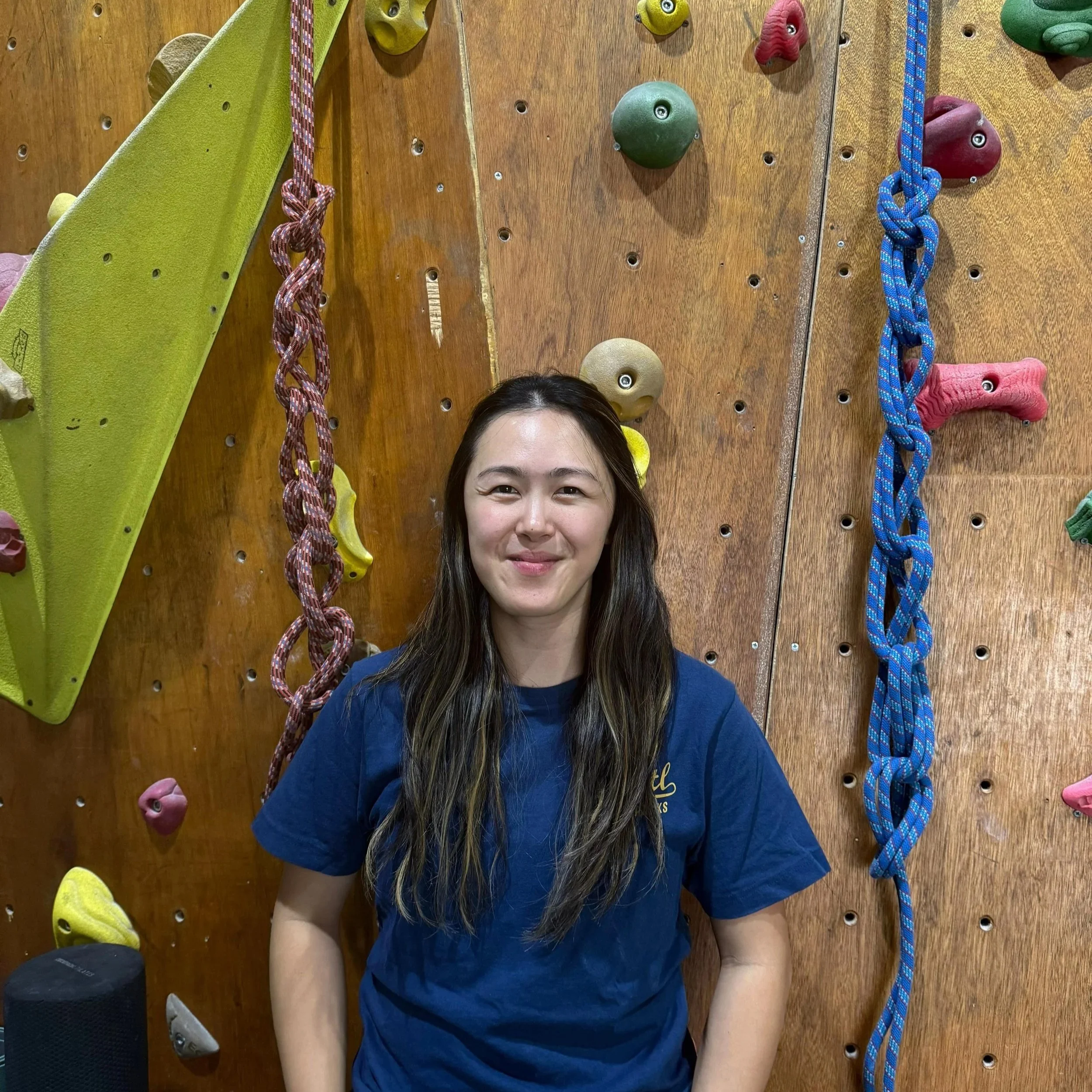 A woman with long dark hair in a blue shirt standing in front of an indoor rock climbing wall with various colorful holds and climbing ropes.