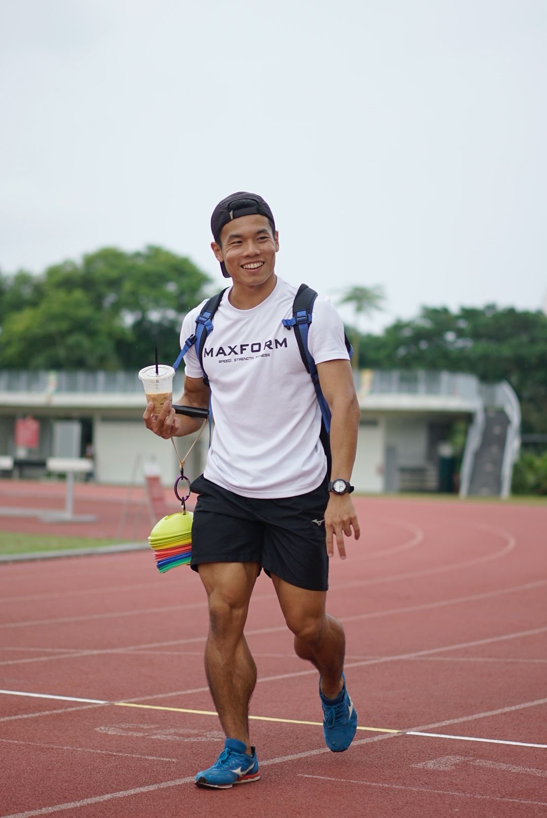 Young man running on an outdoor track, smiling, wearing a white t-shirt, black shorts, blue running shoes, a watch, and a cap backwards, holding a drink and a set of colorful disc-shaped items hanging around his neck.