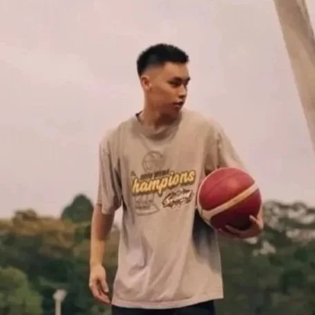 A young man holding a basketball outdoors on a sports field, wearing a beige t-shirt with the word 'champions' printed on it.