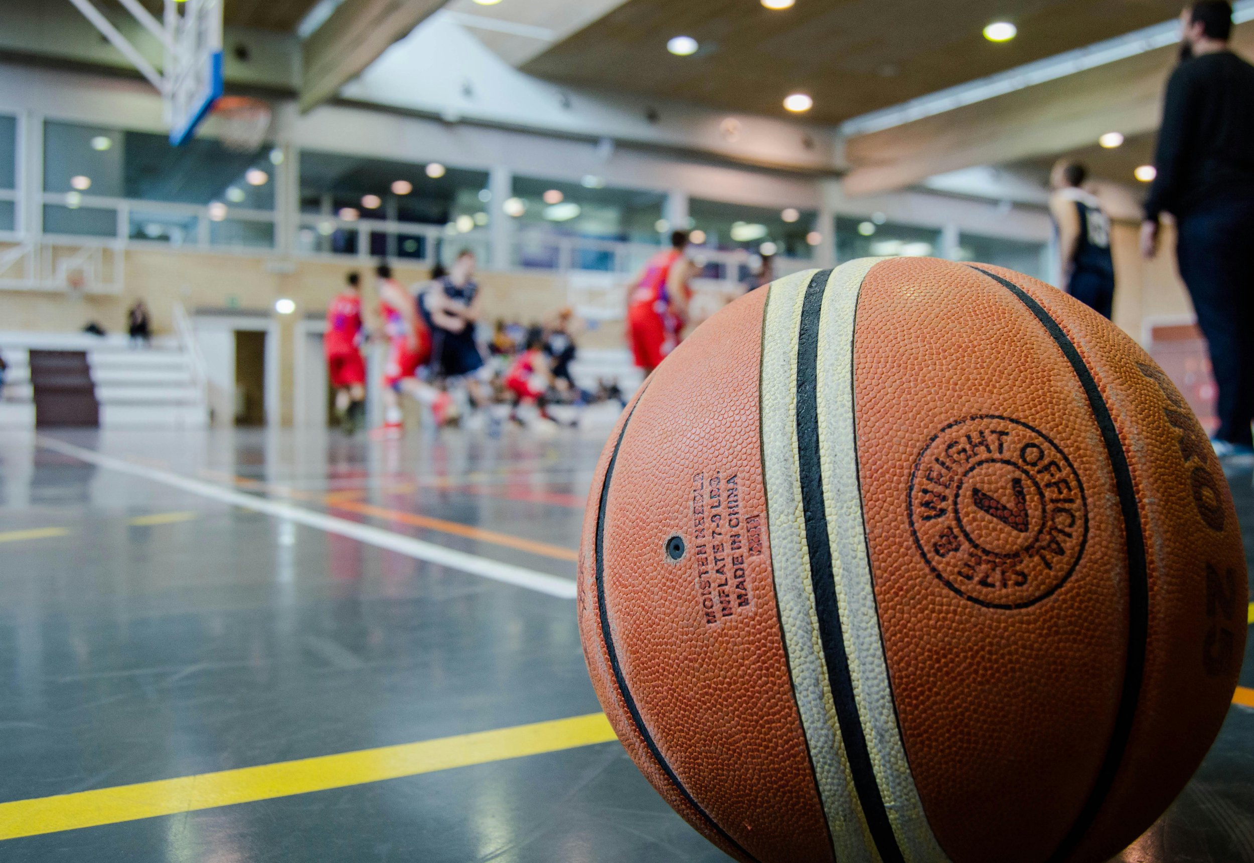 An indoor basketball court with players and coaches, with a close-up of a basketball in the foreground.