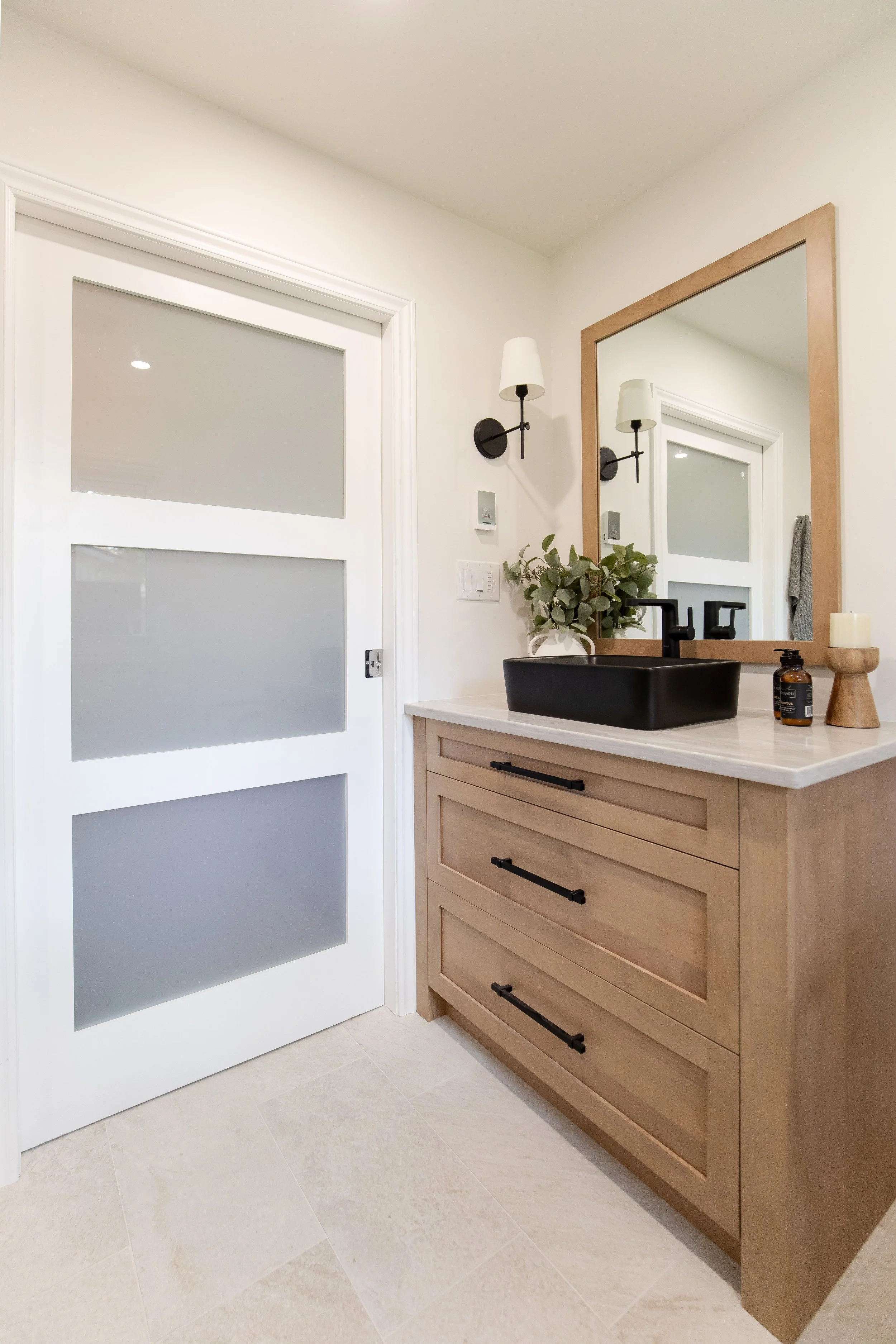 Modern bathroom vanity with a black vessel sink, wooden cabinet with black handles, large mirror, plant, candle, soap dispenser, wall-mounted light fixtures, and frosted glass sliding door.