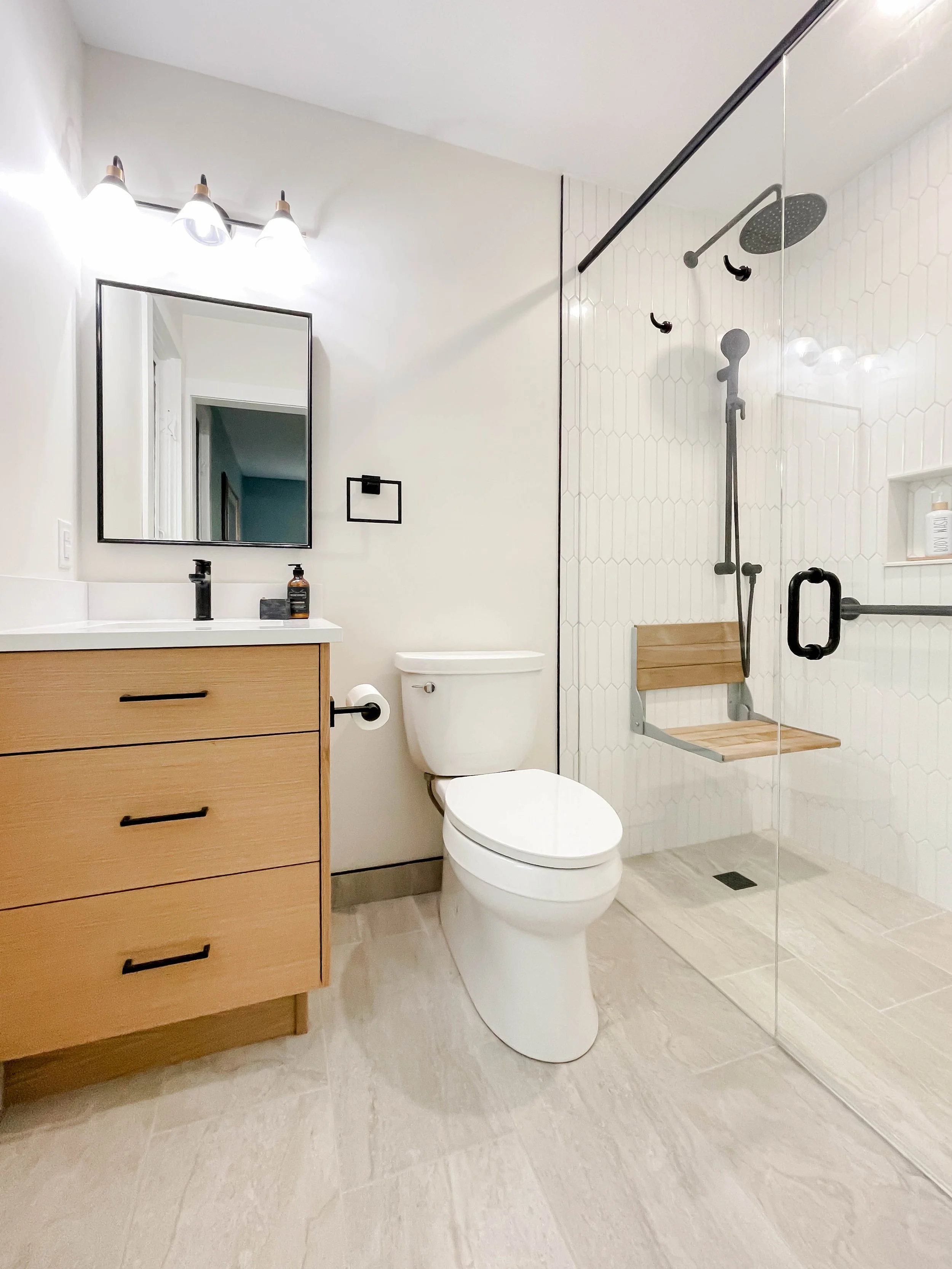 Modern bathroom with wooden vanity, white sink, mirror, toilet, walk-in shower with glass door, wooden bench, and black fixtures.