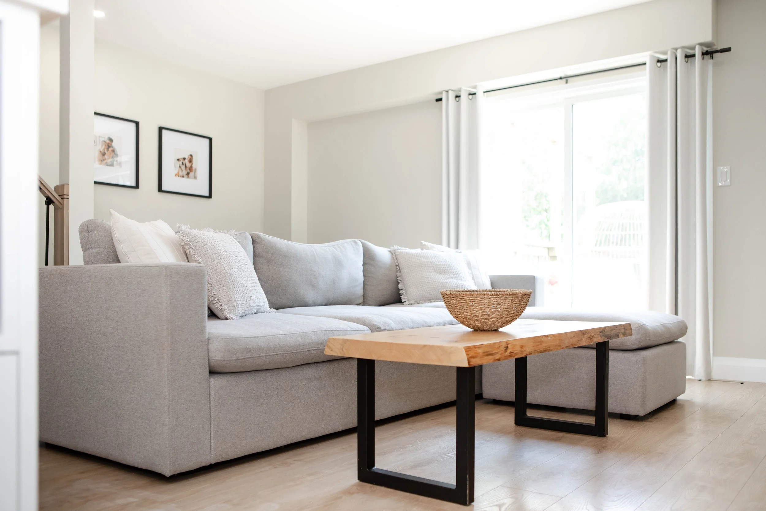 Modern living room with a light gray sectional sofa, wooden coffee table with black metal legs, white curtains, and framed photos on a white wall.