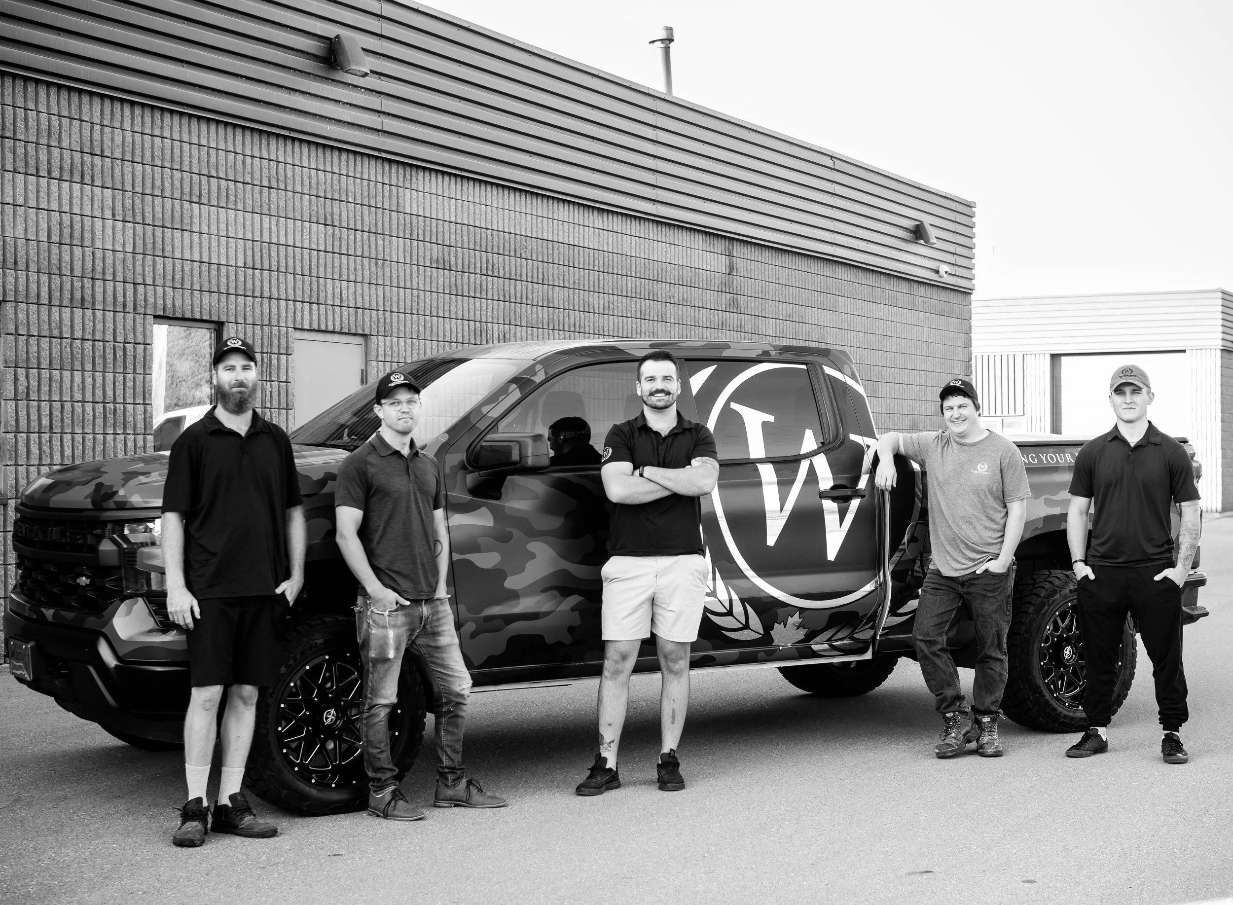 Group of five men standing next to a camouflaged pickup truck with a large 'W' logo on the side, outside a commercial building.