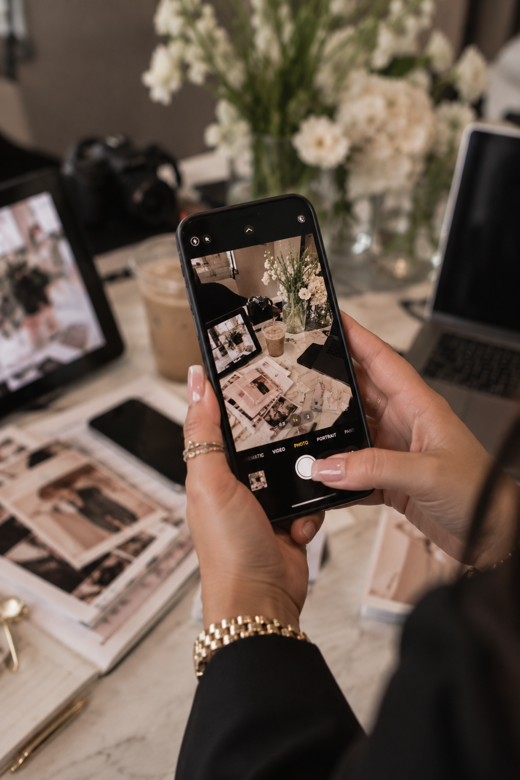Person taking a photo of a table with photo prints, a magazine, a camera, and a coffee drink, with a large vase of white flowers in the background.