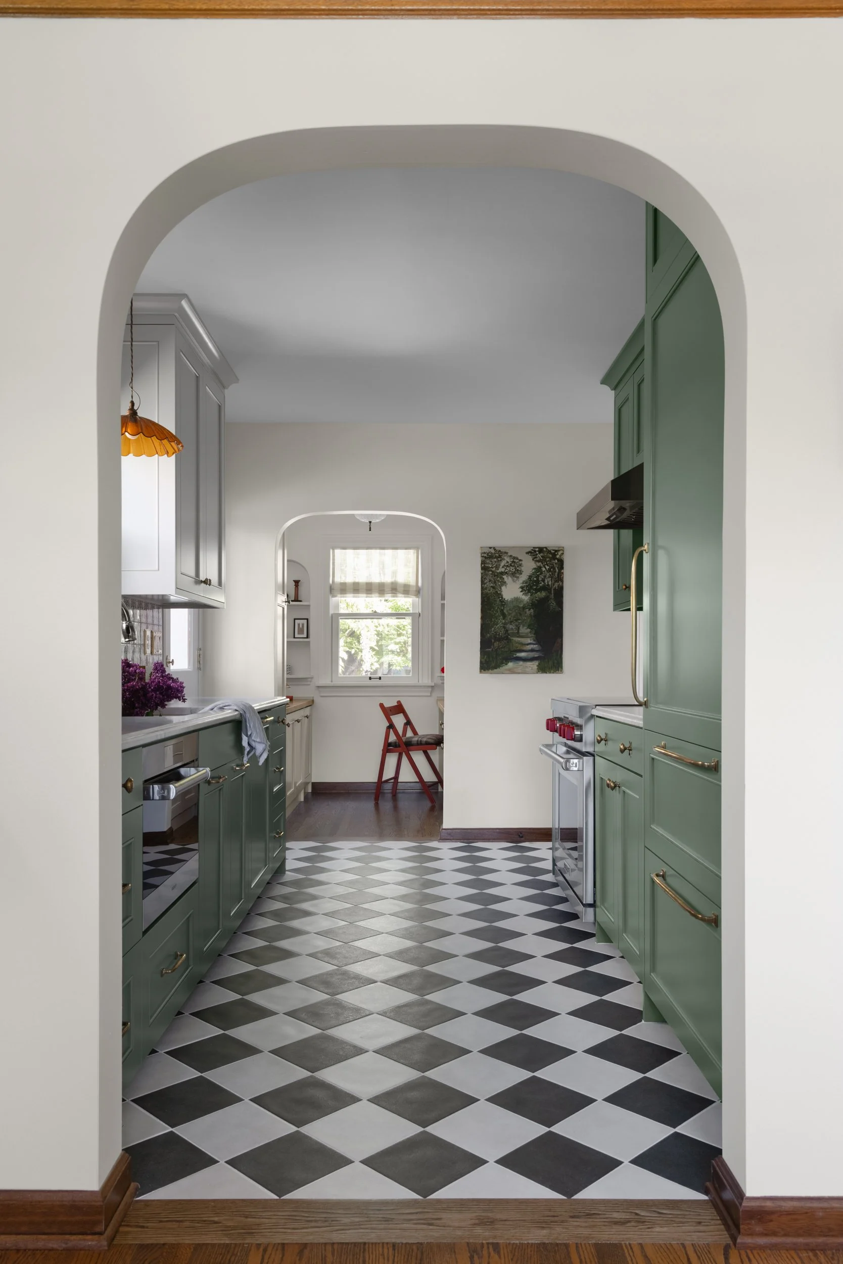 View through a plaster arch highlighting a remodeled kitchen with green cabinets, black-and-white checkerboard flooring and a multifunctional office nook by Full Bloom Interior Design in Seattle.