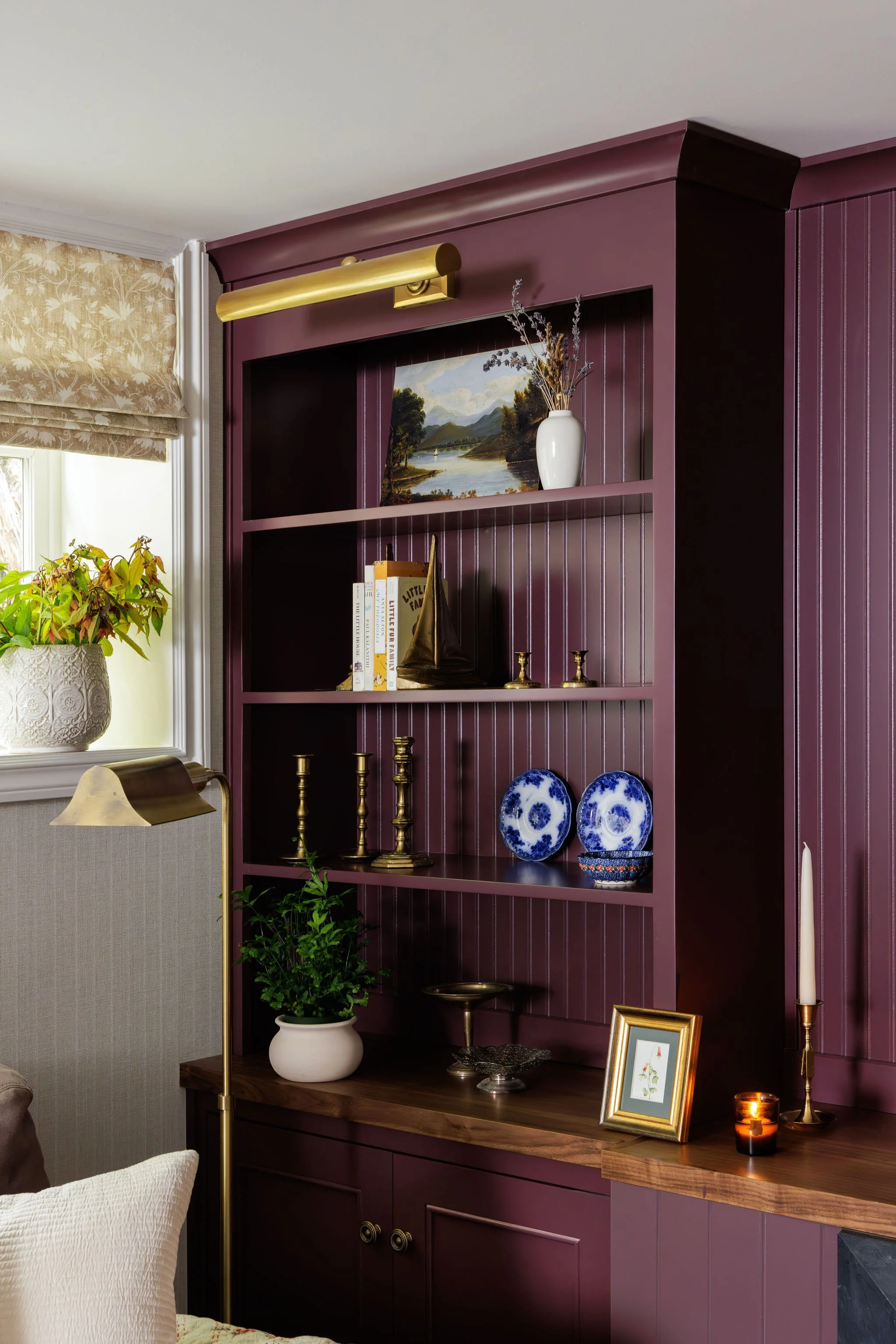 A burgundy built-in shelf with decorative items like vases, books, and candlesticks, next to a window with beige floral shades and a beige armchair.