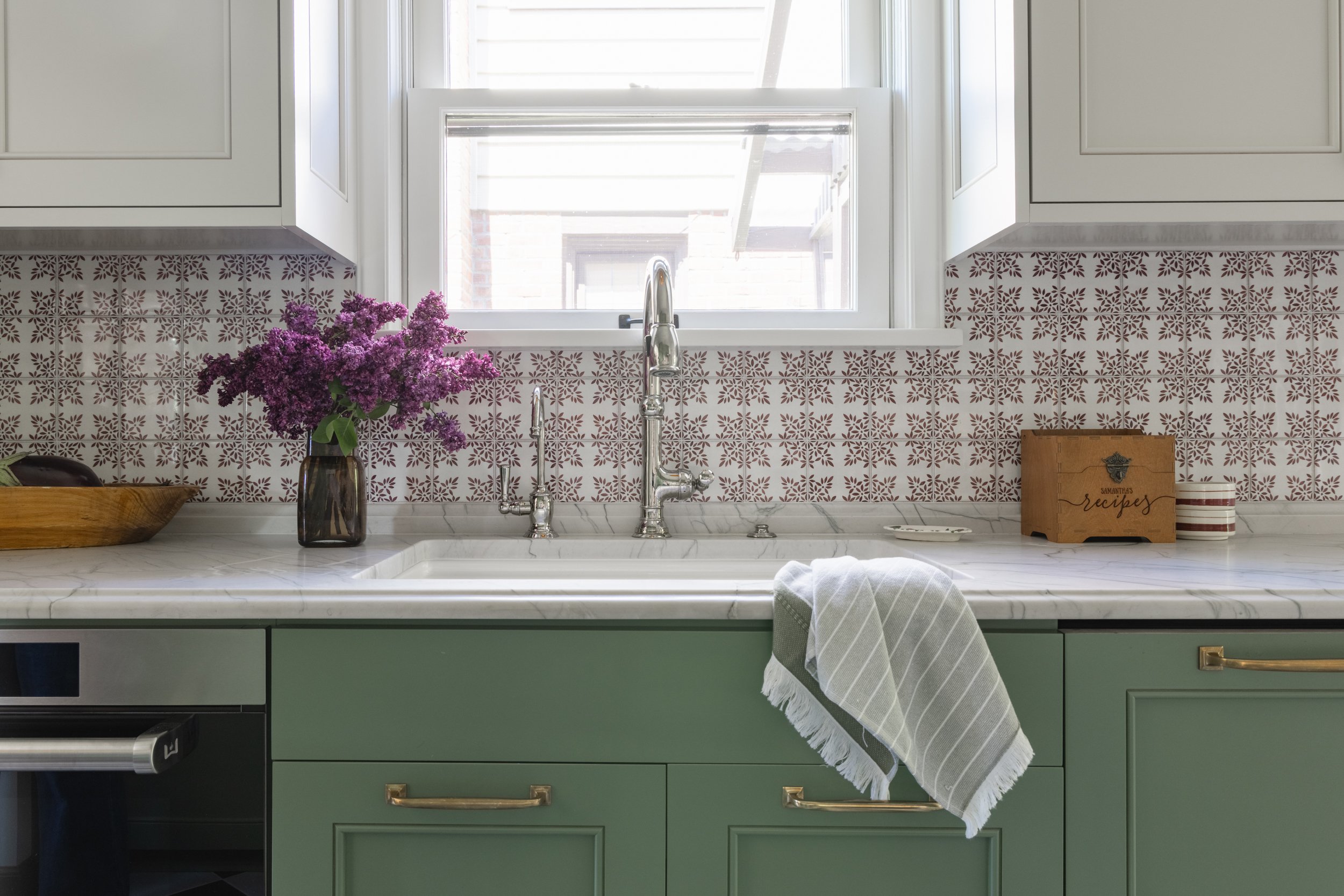 Detailed view of a kitchen sink with muted green cabinetry, patterned tile backsplash, marble countertops, and an antique-style faucet by Full Bloom Interior Design in Seattle.