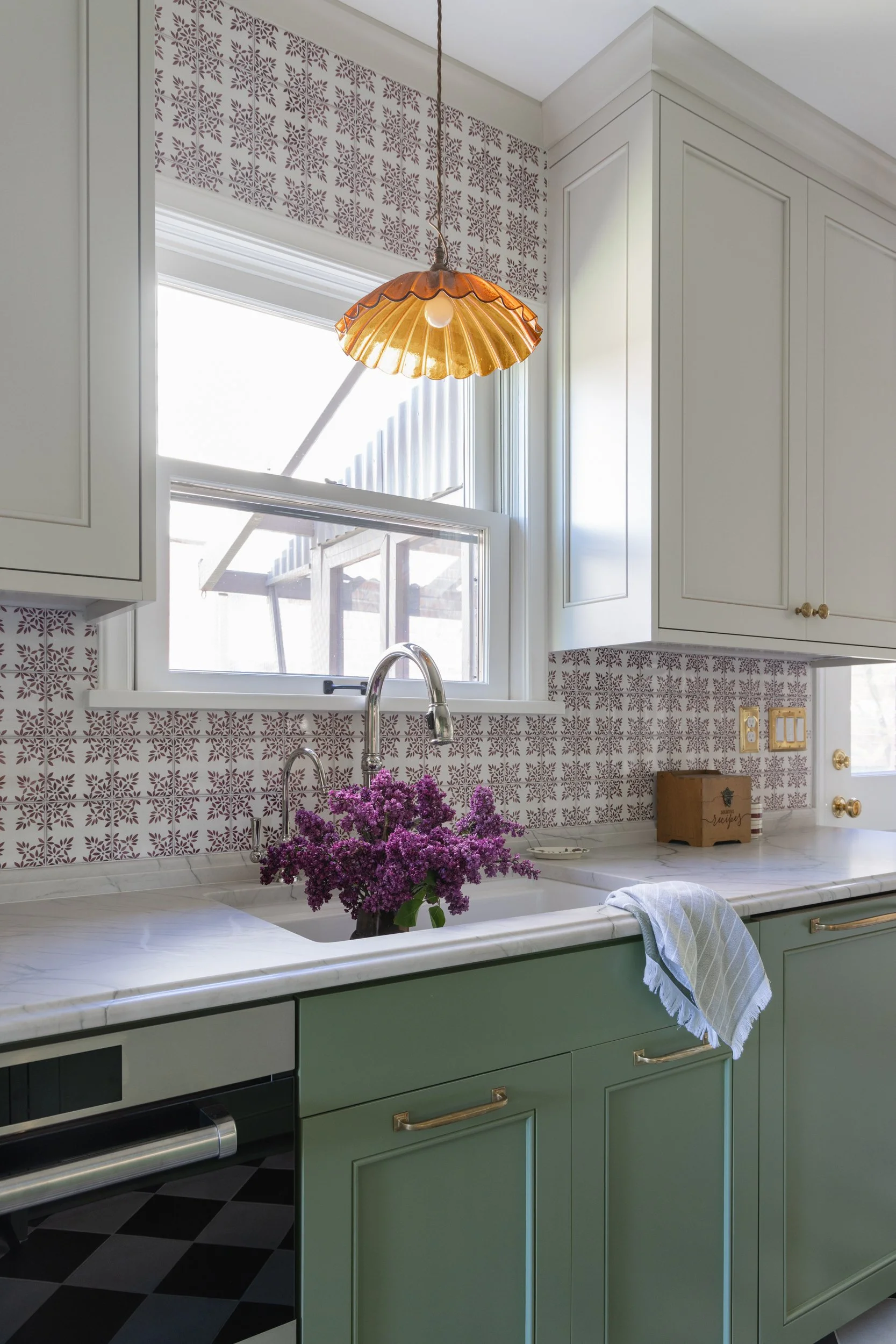 Kitchen sink with marble countertops, muted green cabinets, patterned tile backsplash, a silver faucet, and an amber glass pendant by Full Bloom Interior Design in Seattle.