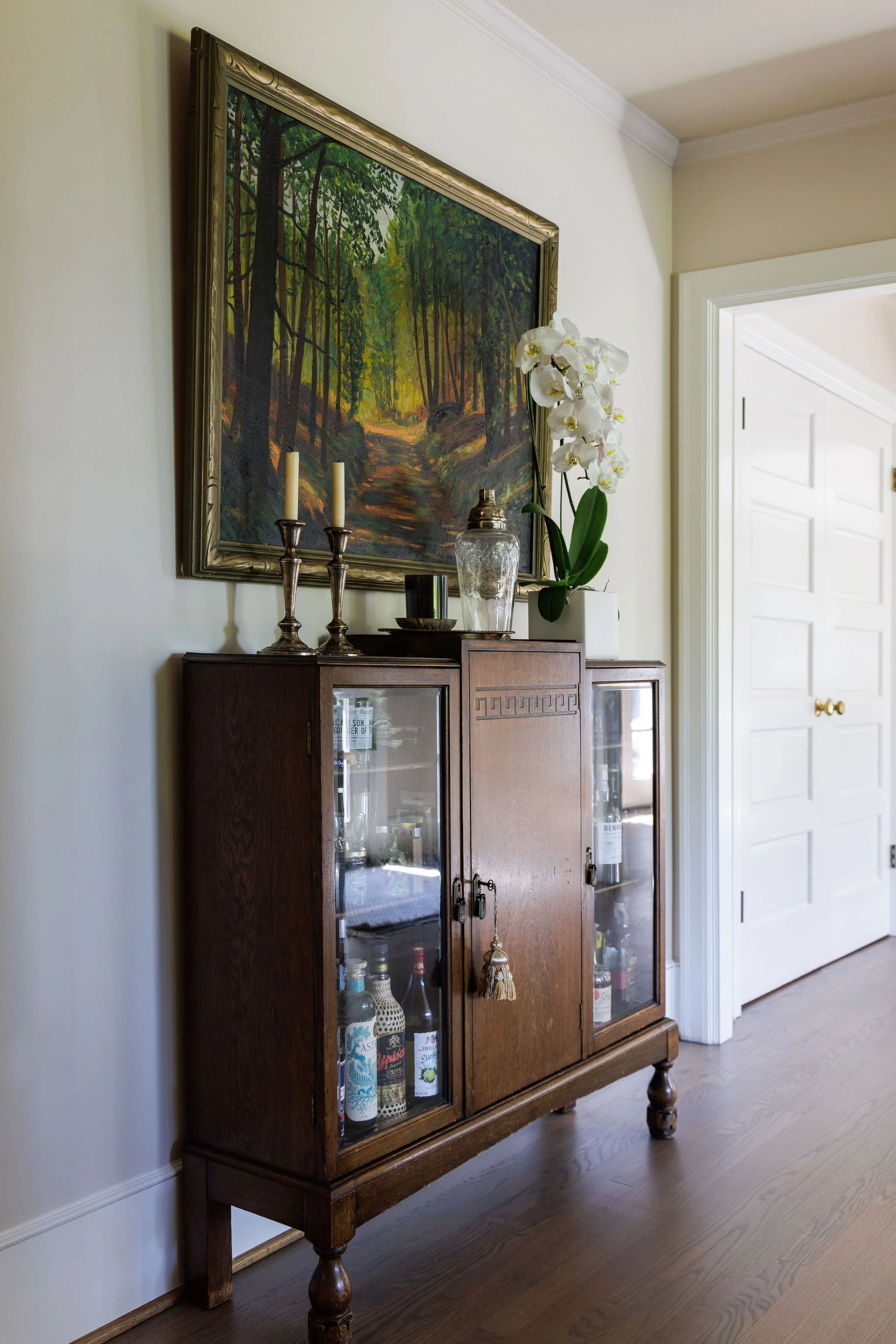Traditional wooden bar cabinet styled with an orchid, landscape oil painting, candlesticks and decor by Full Bloom Interior Design in Seattle.