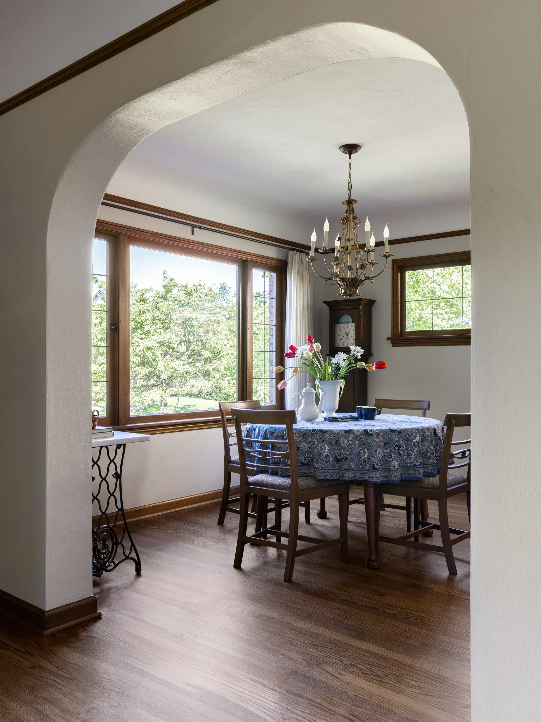 View through a plaster arch into a dining room featuring original wood windows, a vintage chandelier, and warm oak flooring by Full Bloom Interior Design in Seattle.