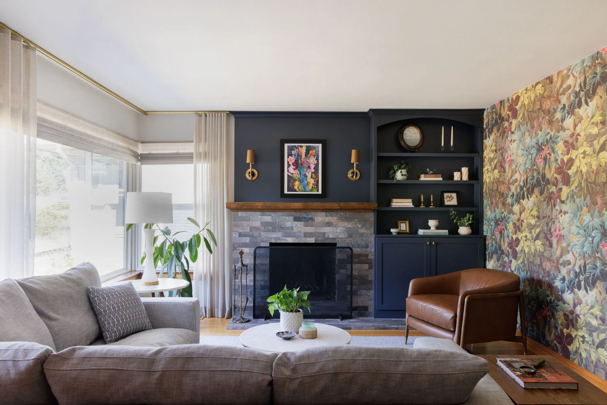 Mid-century modern living room featuring a navy blue accent wall, brick fireplace with a wood mantel, camel leather armchair and colorful floral wallpaper by Full Bloom Interior Design in Seattle.