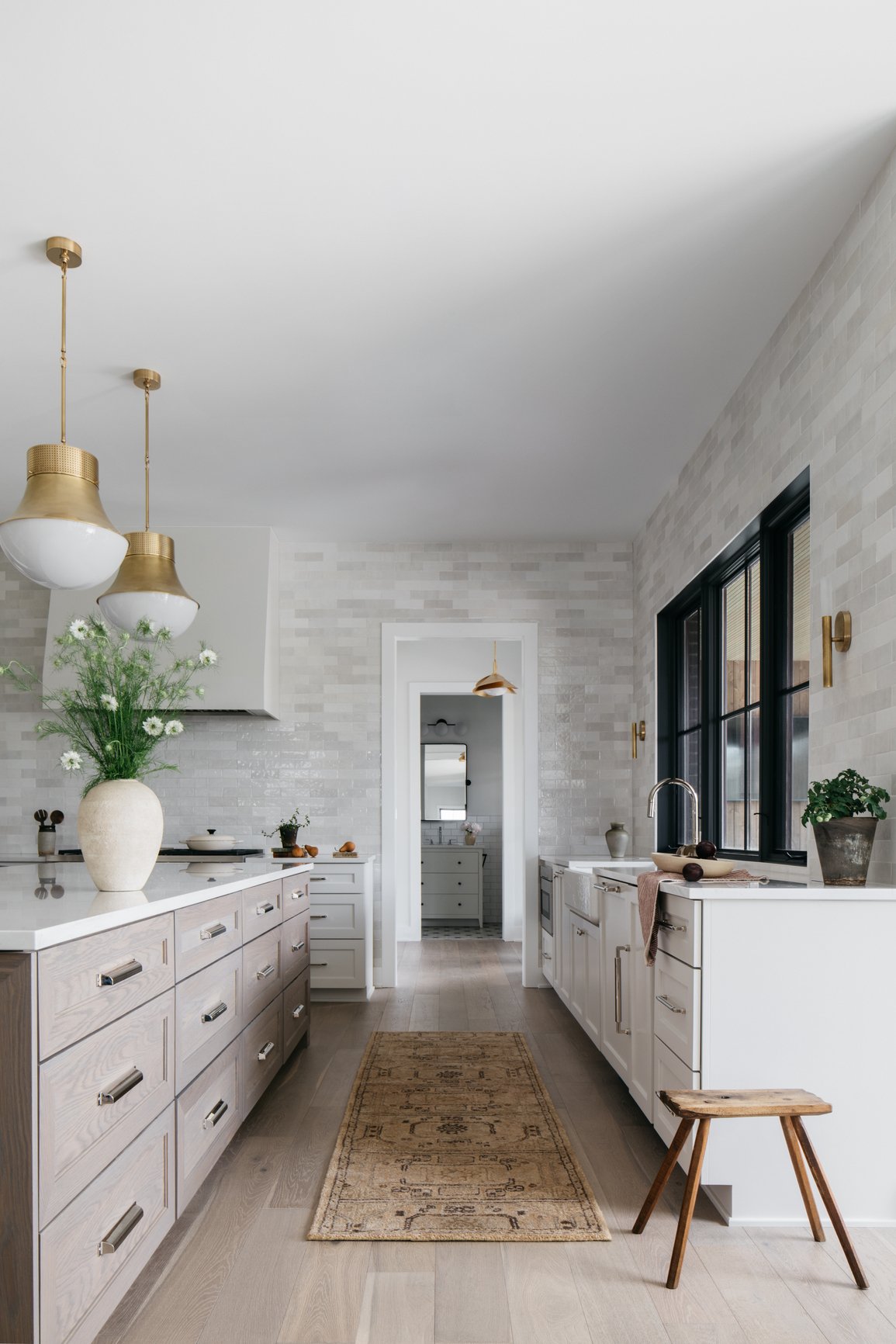 Transitional kitchen with white cabinets, black-framed windows, gold light fixtures, potted plants, and wooden stool on a light wood floor.